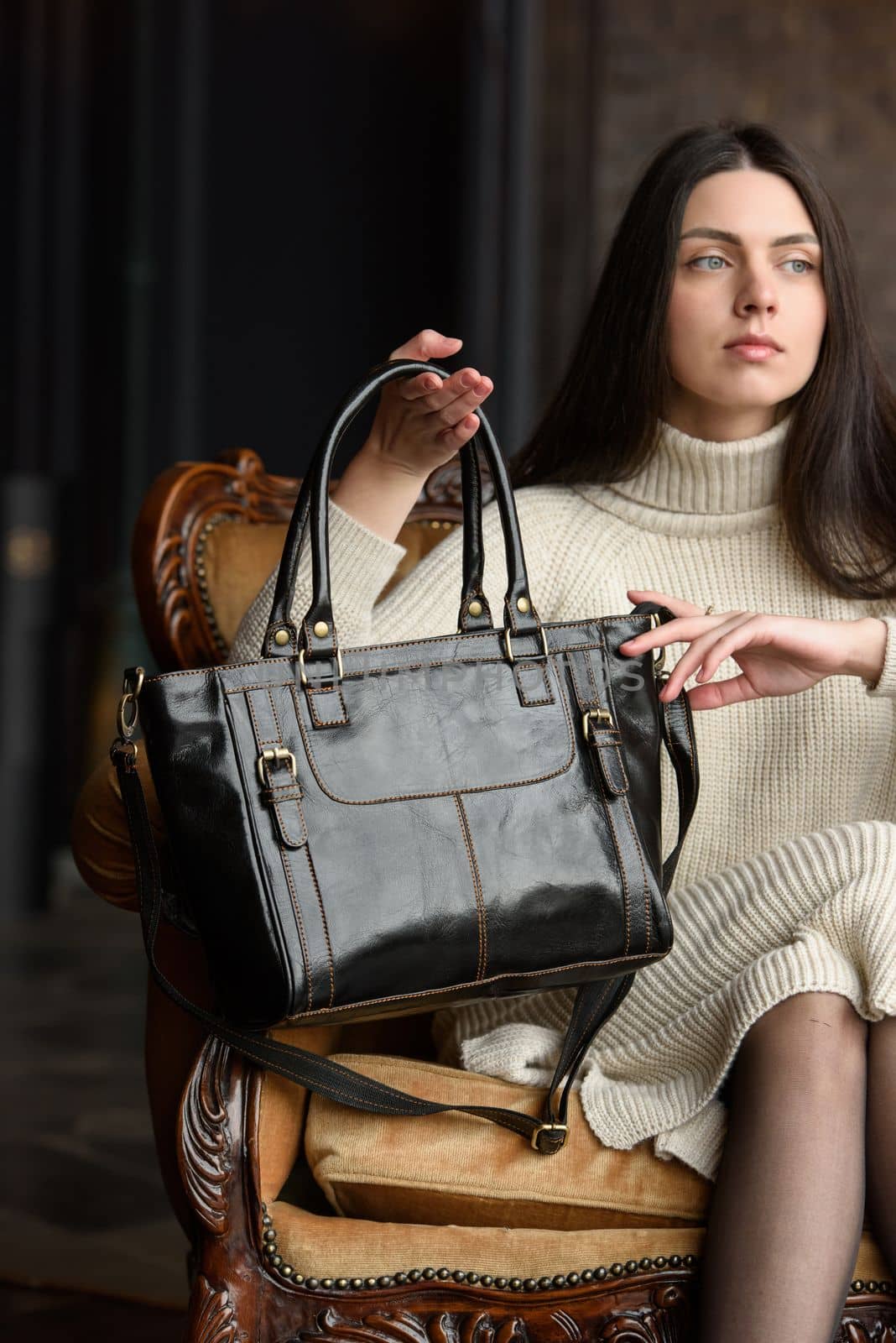 a brunette girl in a knitted beige dress poses while sitting with a shiny black leather bag in her hands