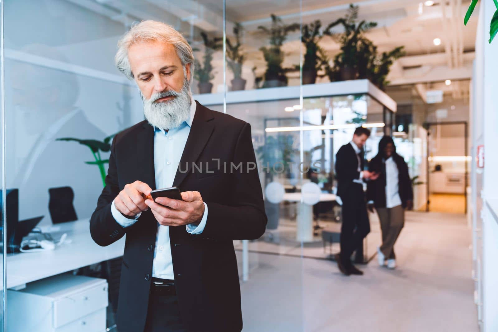 Senior business man with grey hair and a beard looking down at his phone, writing an email by VisualProductions