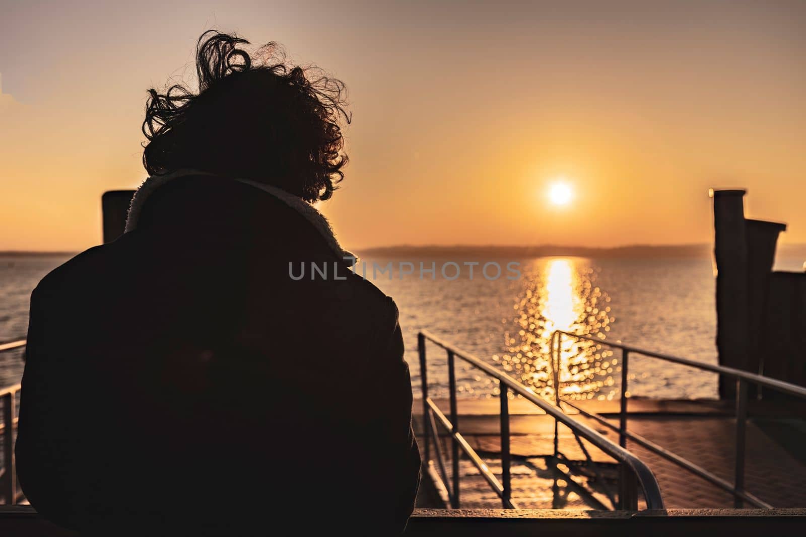 A young boy with his back turned, gazing thoughtfully at the sunset over a tranquil lake