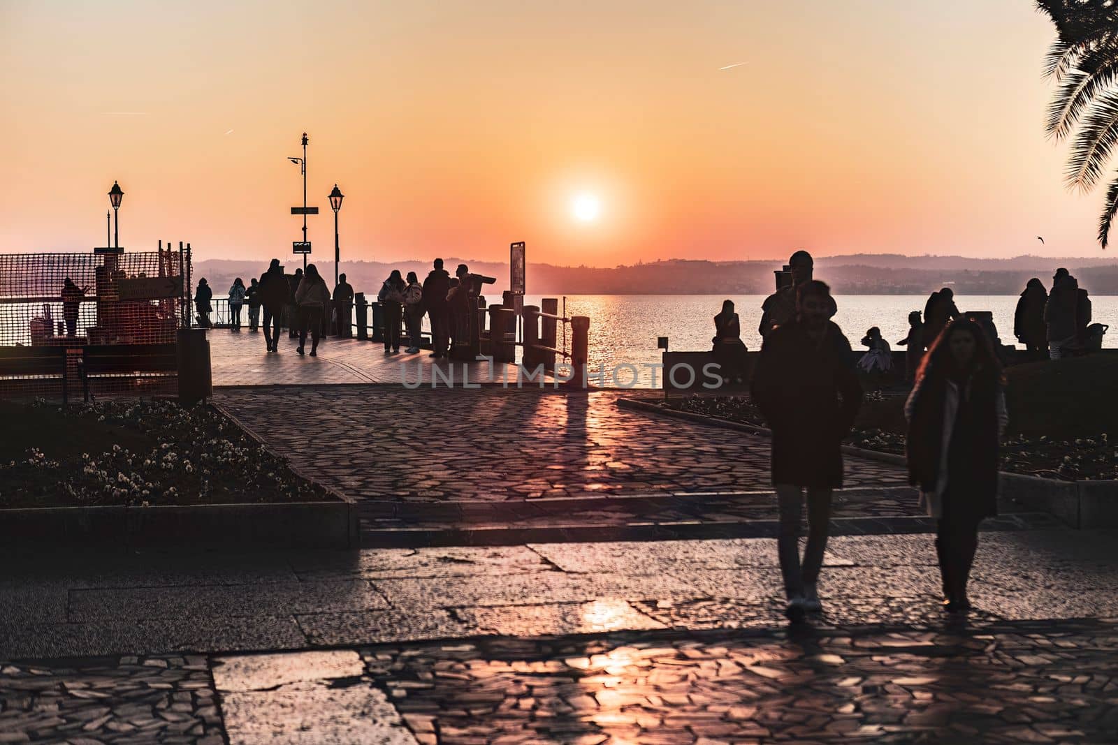 Sirmione, Italy 15 February 2023: A stunning view of Sirmione's landscape at sunset with orange hues, silhouetted people, and the serene lake in the background.