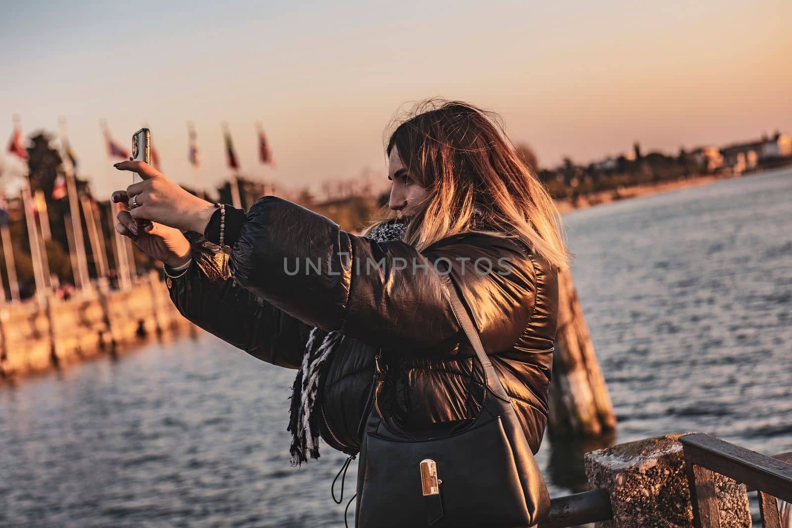 Sirmione, Italy 15 February 2023: A young woman takes a selfie on the pier, capturing the beautiful sunset behind her. She looks happy and content in this scenic moment