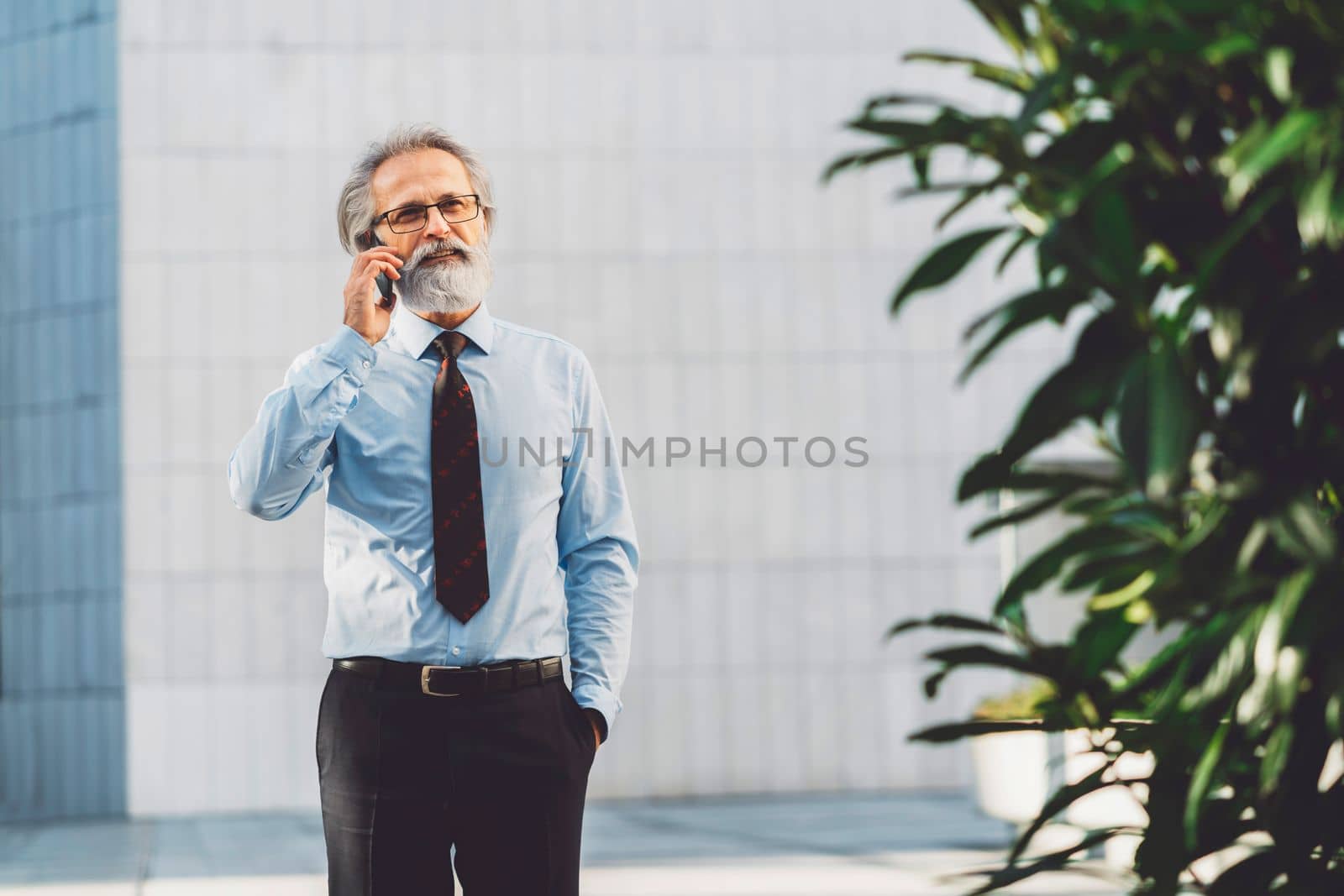 Senior business man with grey hair and a beard talking on the phone outside on a sunny day. CEO on the phone call outside.