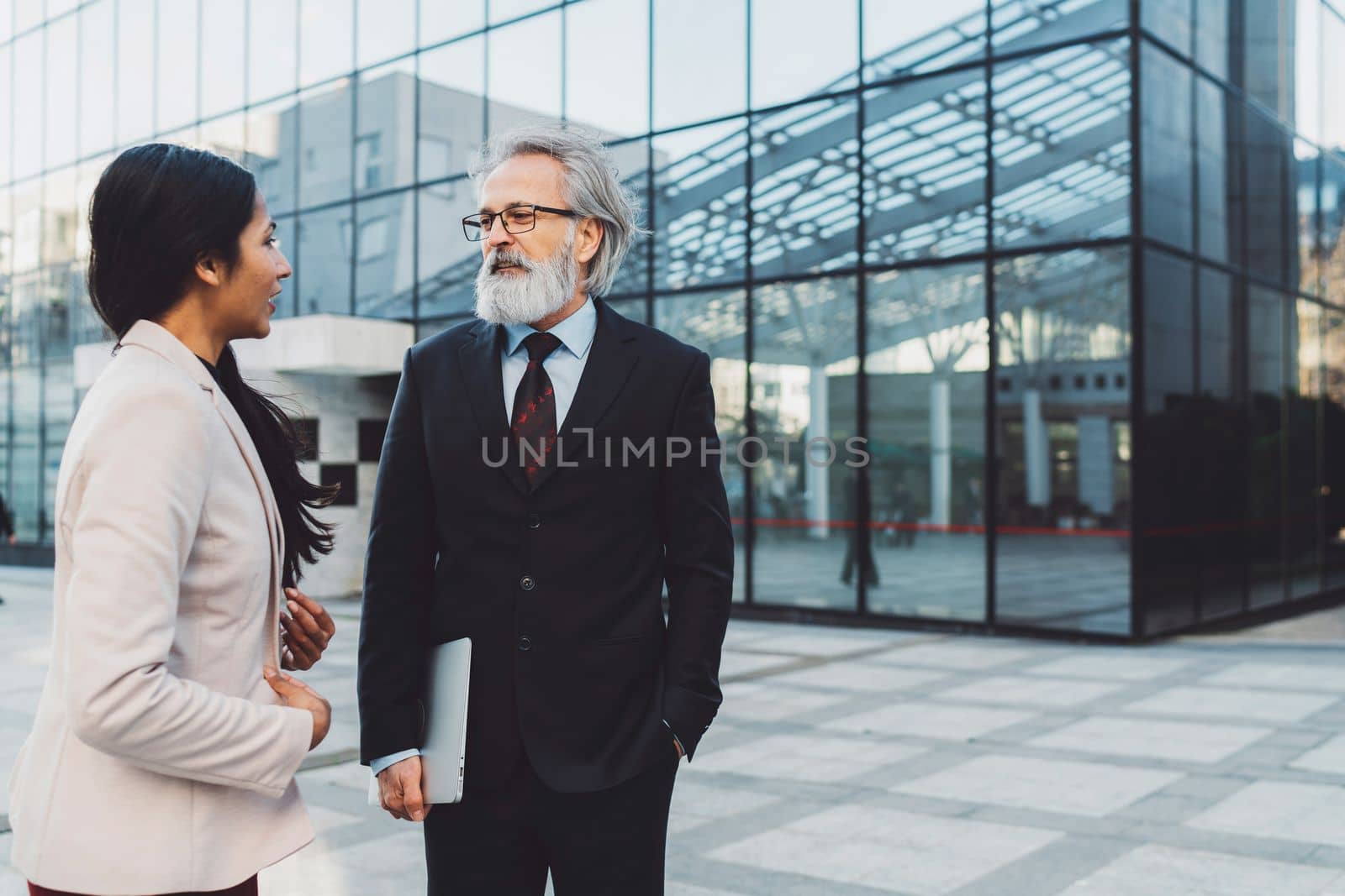Business man and business woman standing outside talking on a sunny day, office building in the background.