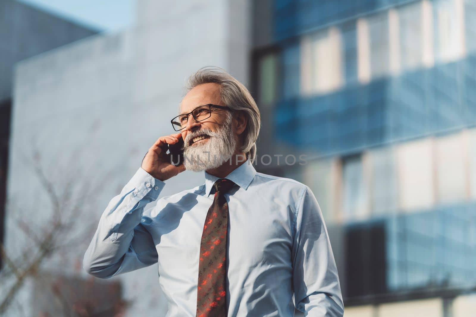 Senior business man with grey hair and a beard talking on the phone outside on a sunny day. CEO on the phone call outside.