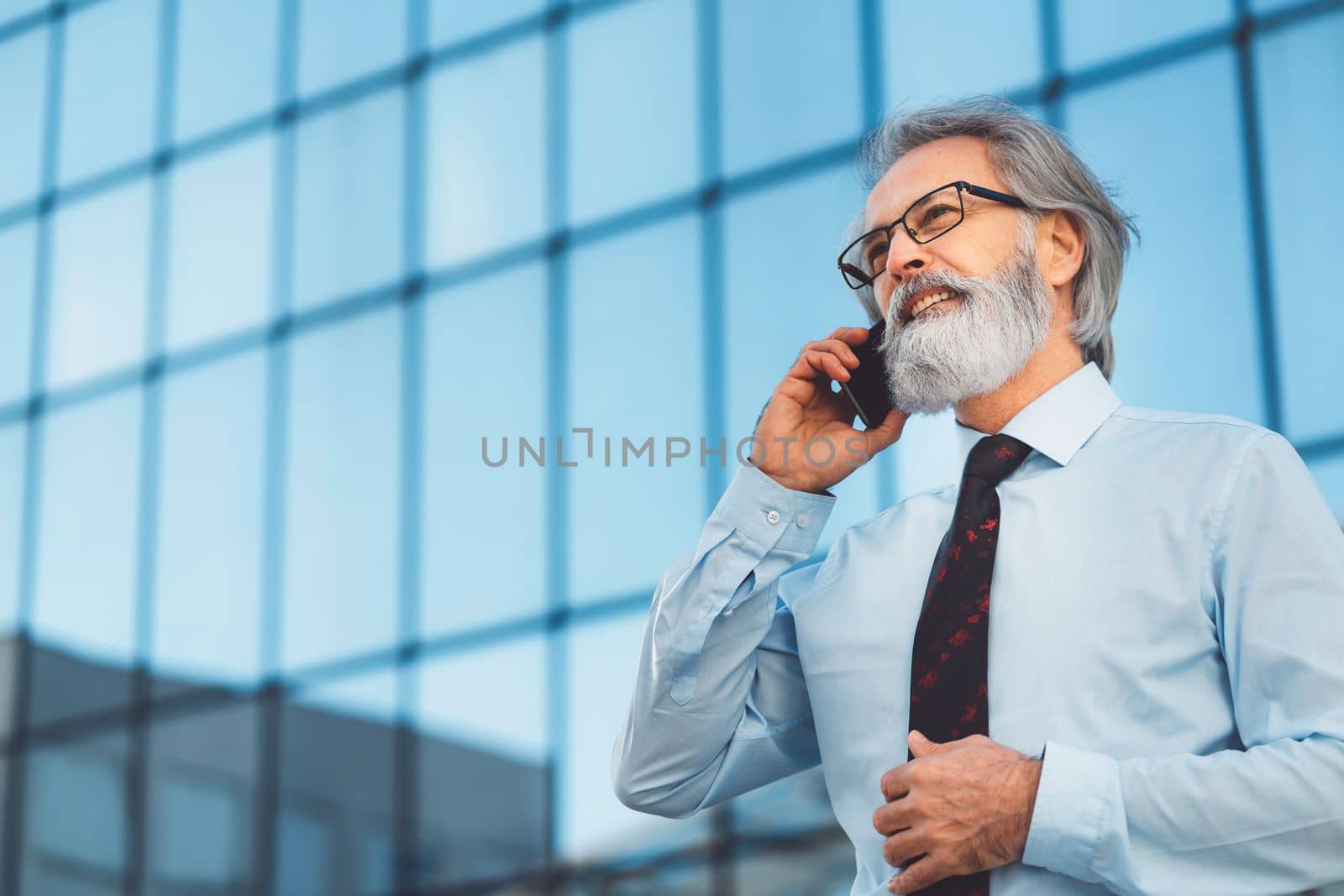 Senior business man with grey hair and a beard talking on the phone outside on a sunny day. CEO on the phone call outside.