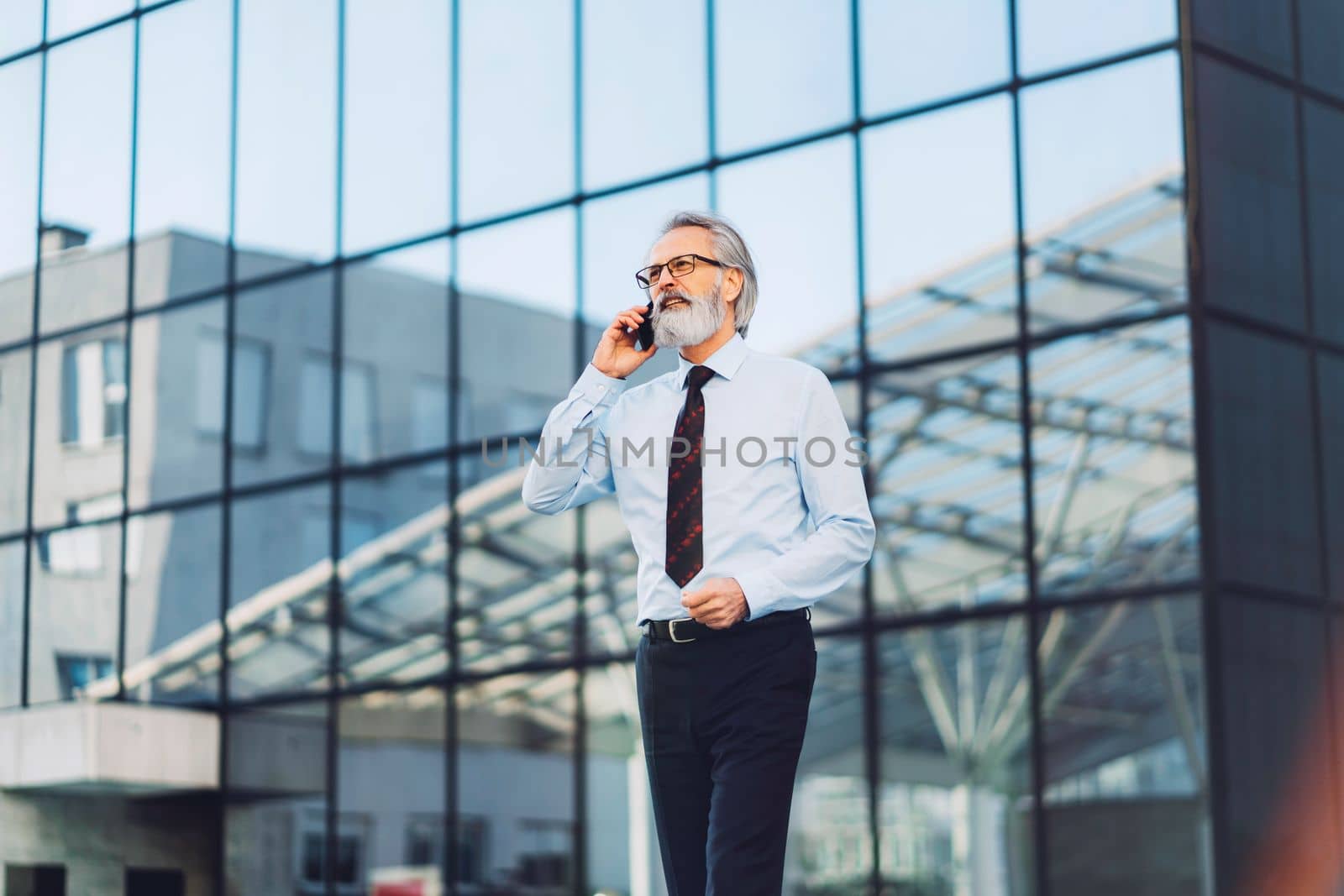 Senior businessman walking outside, glass building in the background, having a phone call by VisualProductions