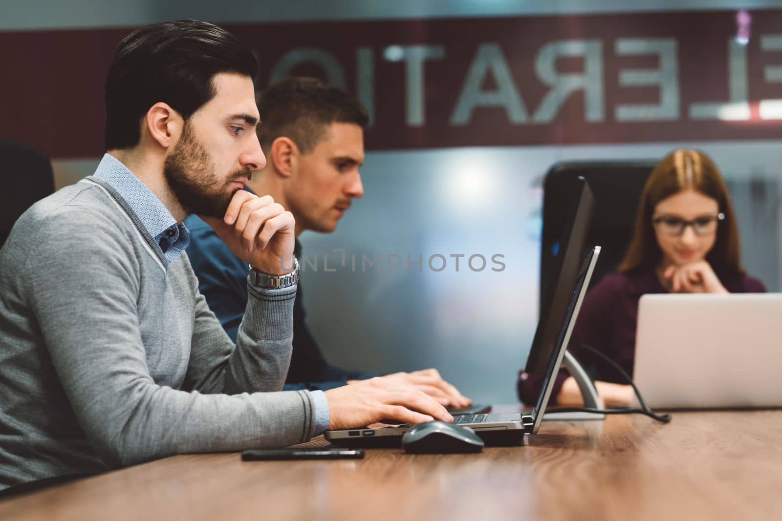 Sitting in front, a young business man working on his laptop, and two of their colleagues, blurred in the background by VisualProductions