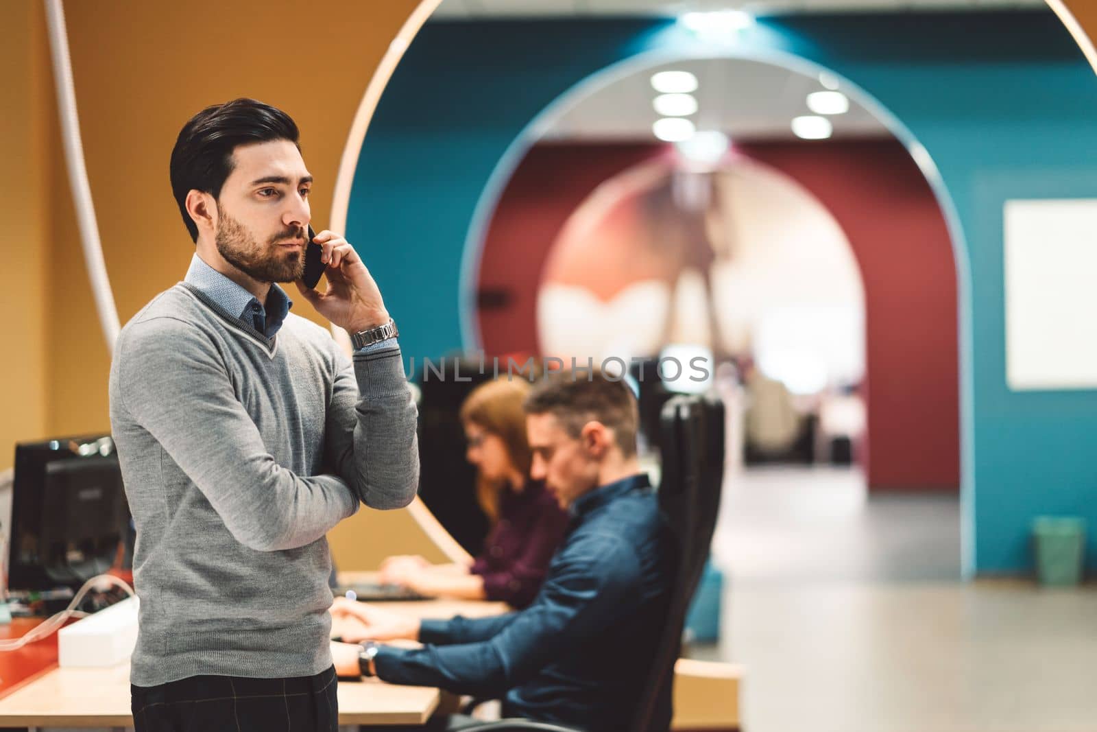 Young bearded man leaning on the office desk while talking on the phone by VisualProductions