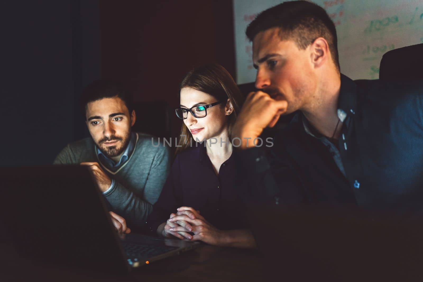 Three business people, two man and a women working in the dark, laptop screen lighting up their faces by VisualProductions