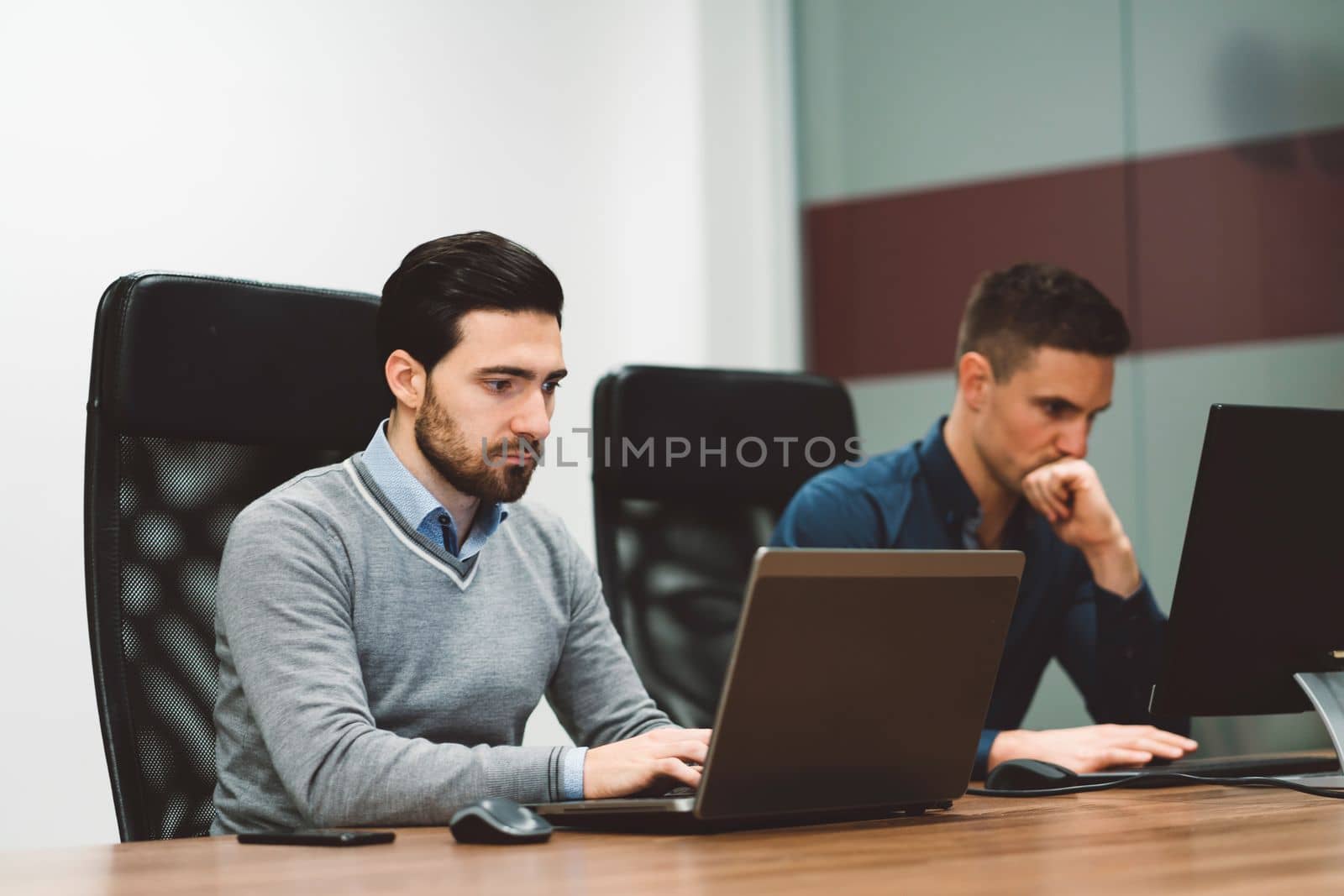 Couple of business man sitting by the desk working on their laptop and computer, with a focused face expression by VisualProductions