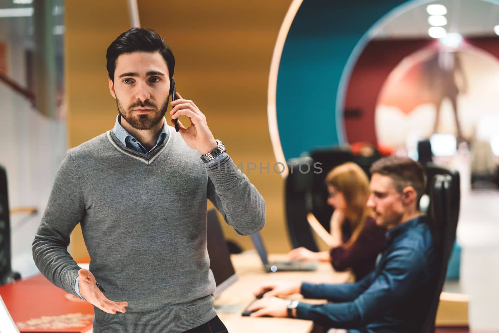 Young caucasian man with beard on a phone call while at the office, blurred in the background people working at their desk by VisualProductions