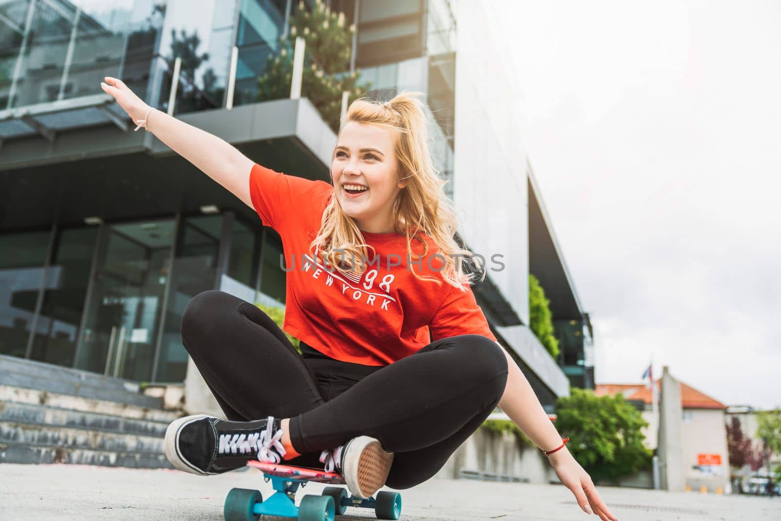 Young blonde caucasian girl having fun outside on a skateboard, wearing black leggings and red shirt, sun flare. Teen girl laughing, having fun outside in the summer.