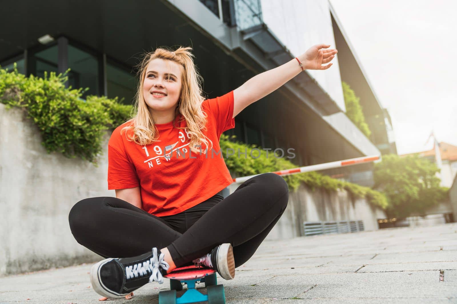 Young blonde caucasian girl having fun outside on a skateboard, wearing black leggings and red shirt, sun flare. Teen girl laughing, having fun outside in the summer.