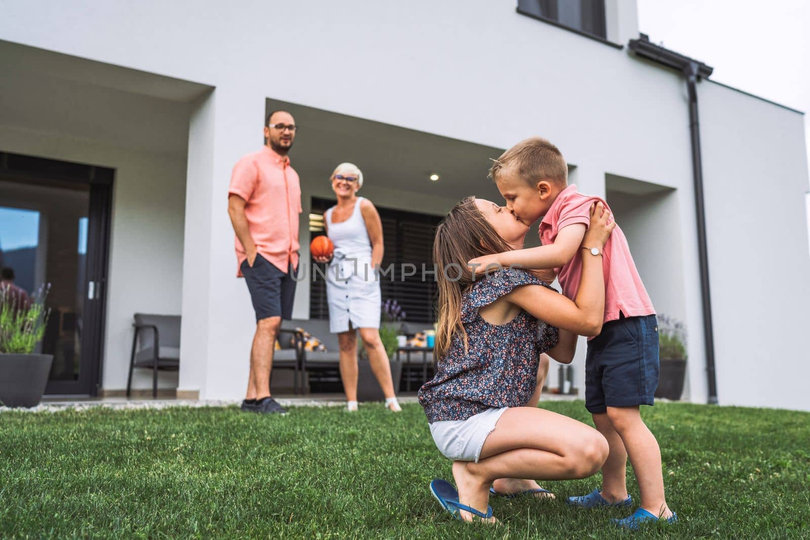 Two young kids, brother and sister kissing while mom and dad are standing in the background - Family of four by VisualProductions