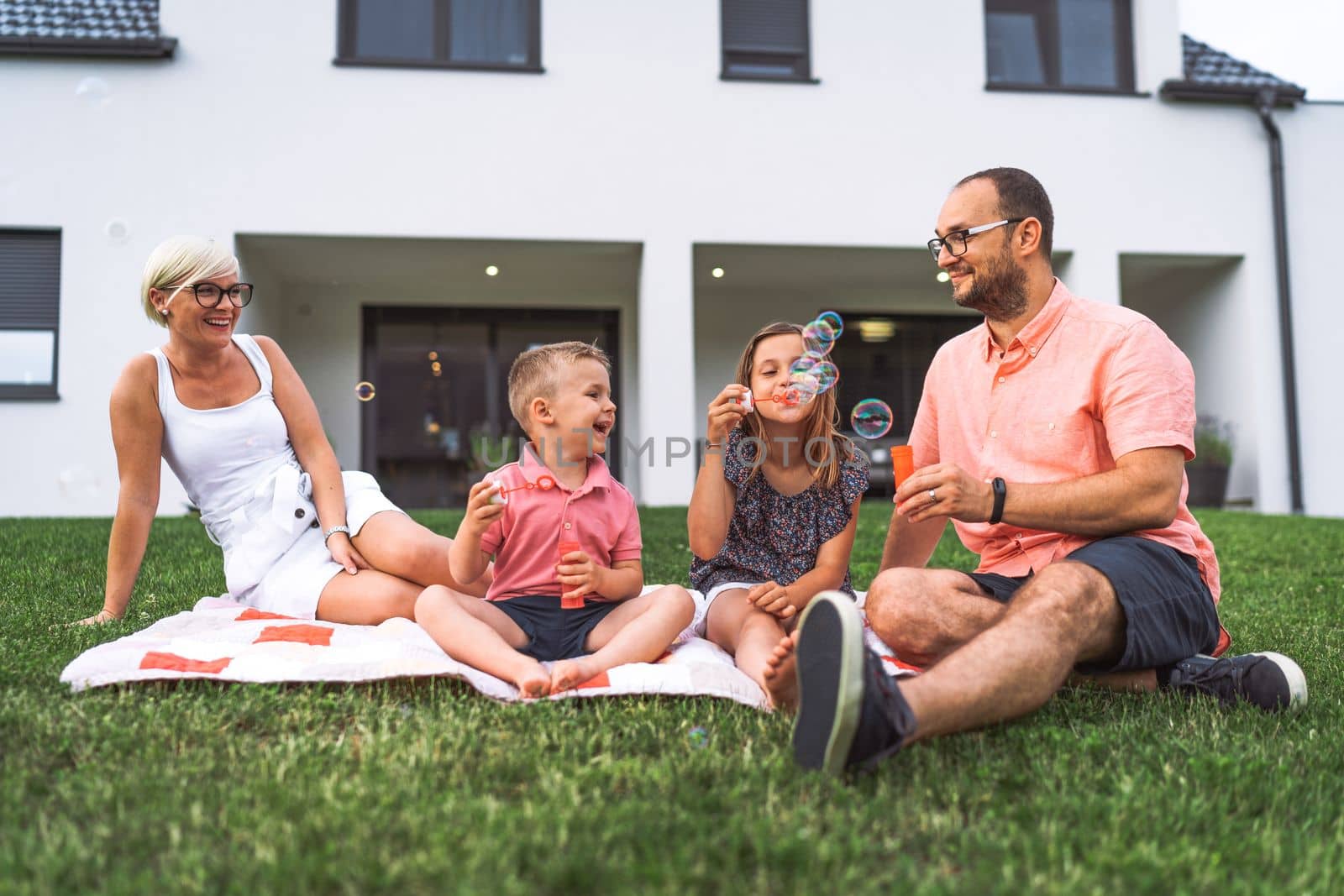 Young caucasian family of four having a picnic outside their house, sitting on a blanket on the grass by VisualProductions