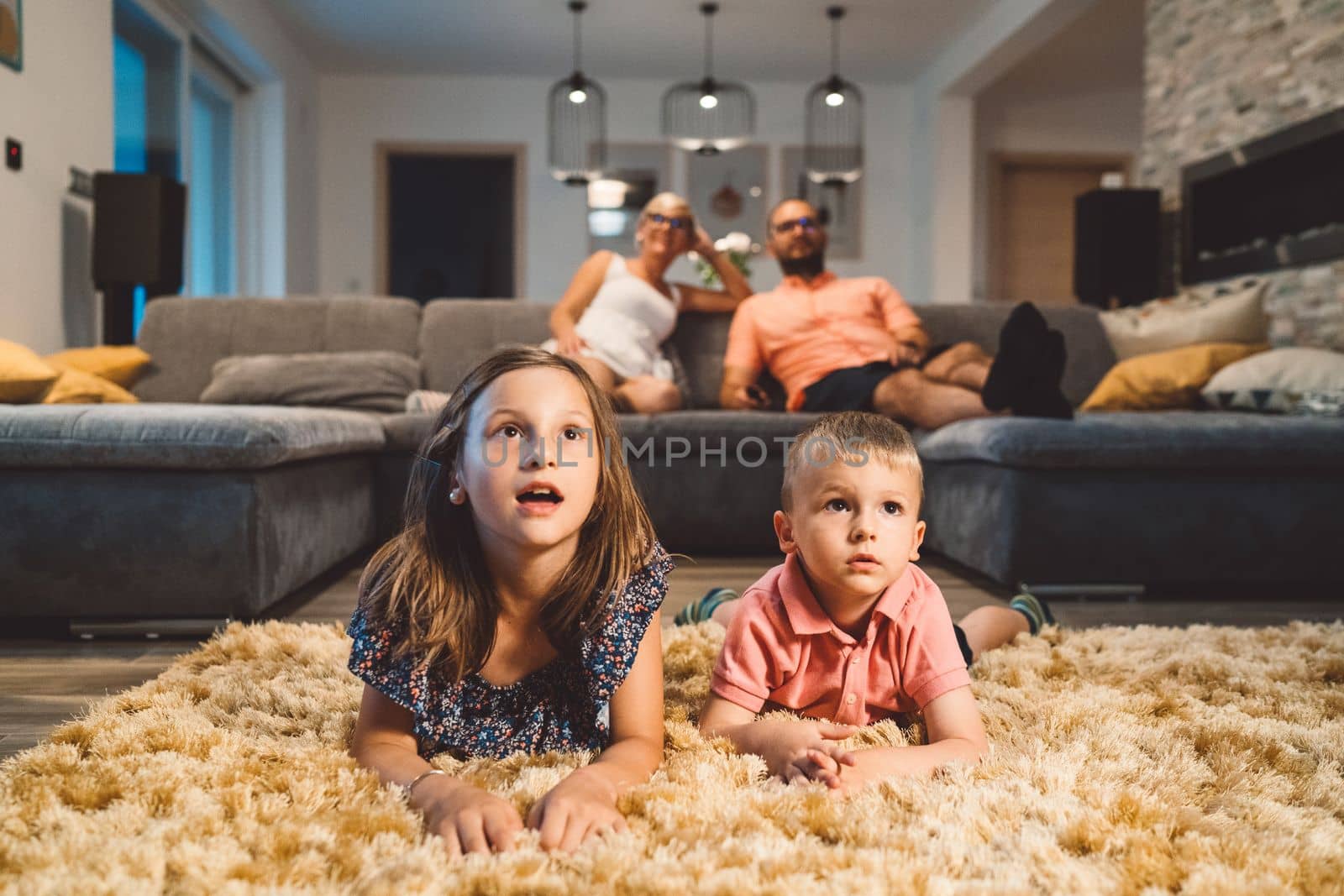 Two young kids laying on the fluffy carpet watching a movie with their parents sitting on the sofa in the background by VisualProductions