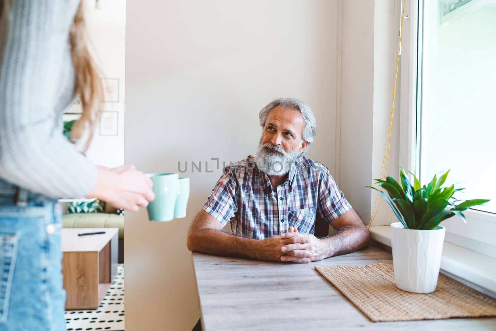 Senior man with grey hair and a beard sitting by the dining table while unrecognizable woman brings coffee to the table by VisualProductions