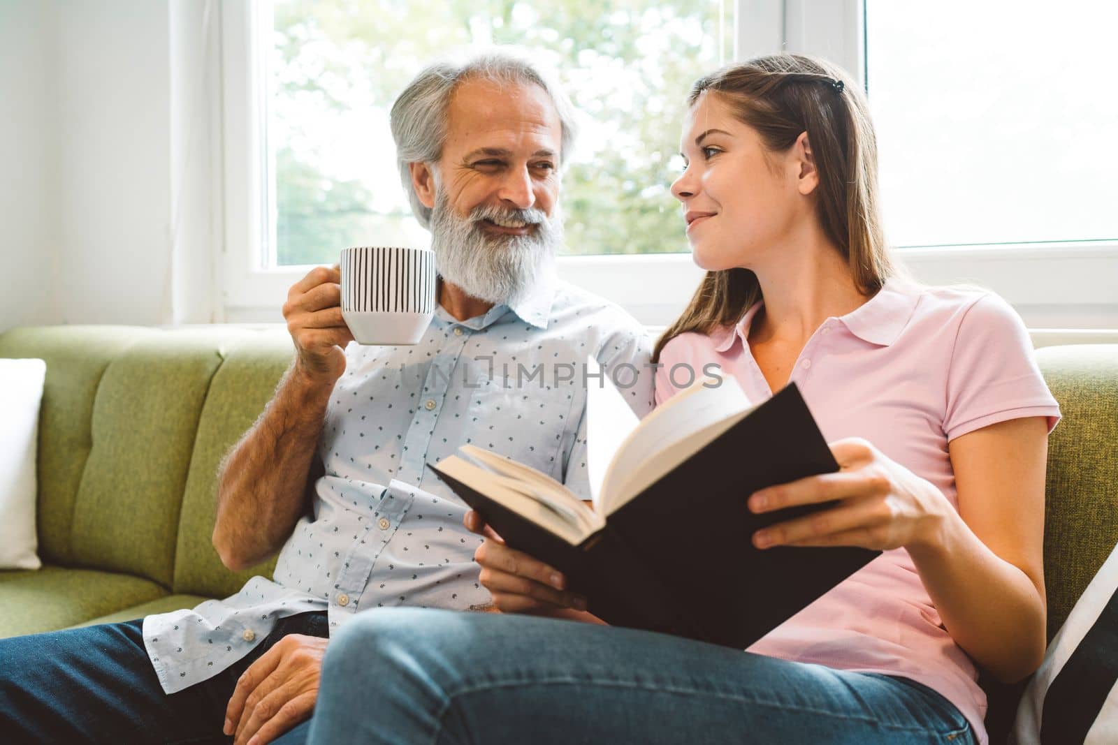 Smiling senior man enjoys spending his time together with his granddaughter, young woman sitting next to him holding a book by VisualProductions