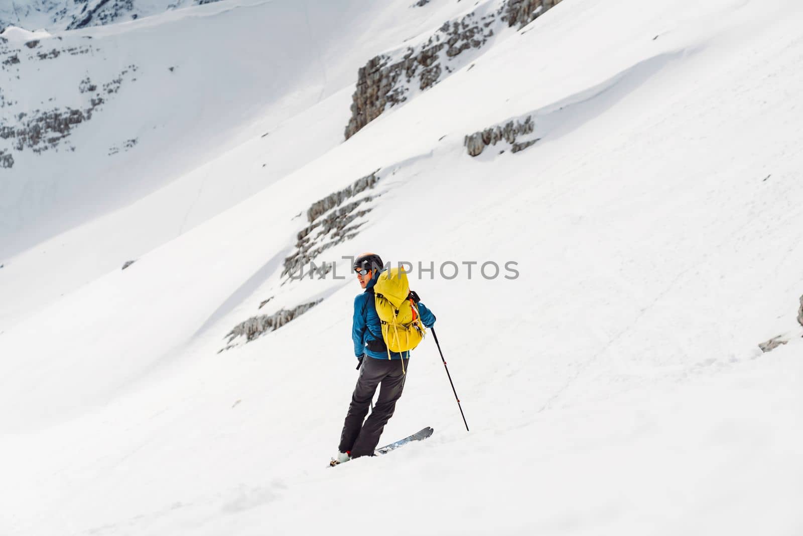 View from the back man ski touring with a yellow backpack, descending down the snowy mountain by VisualProductions
