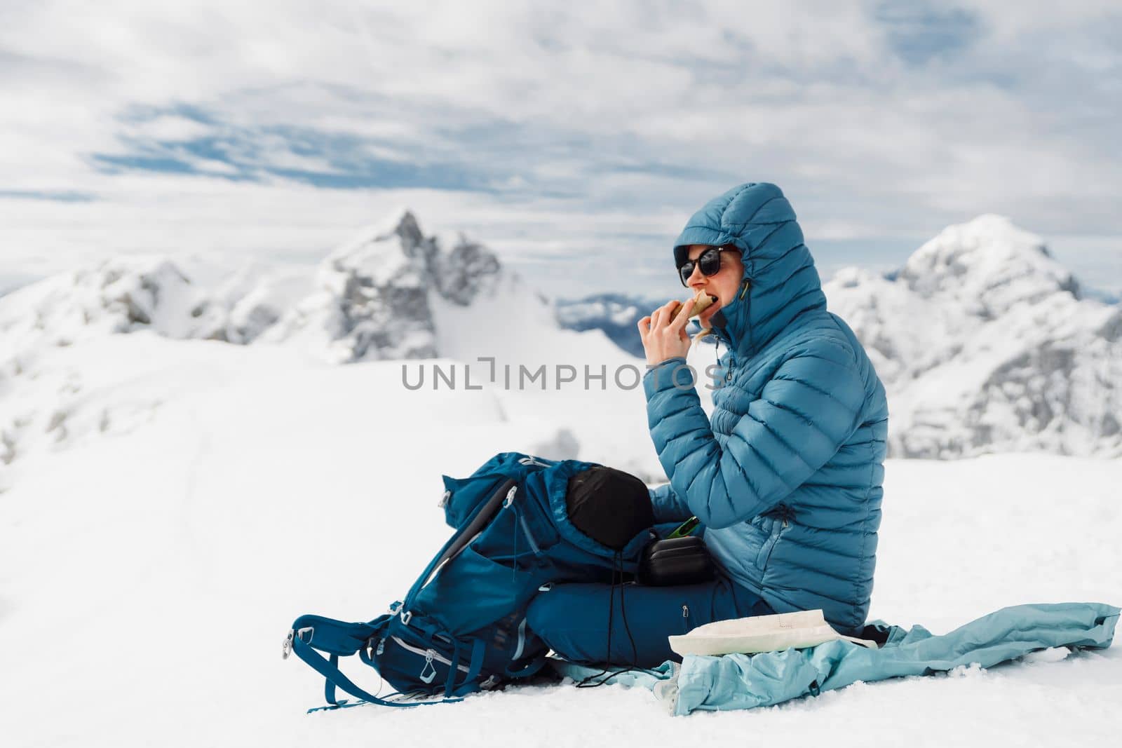 Mountaineer in blue jacket sitting on cold snow eating a sandwich on top of the mountain by VisualProductions