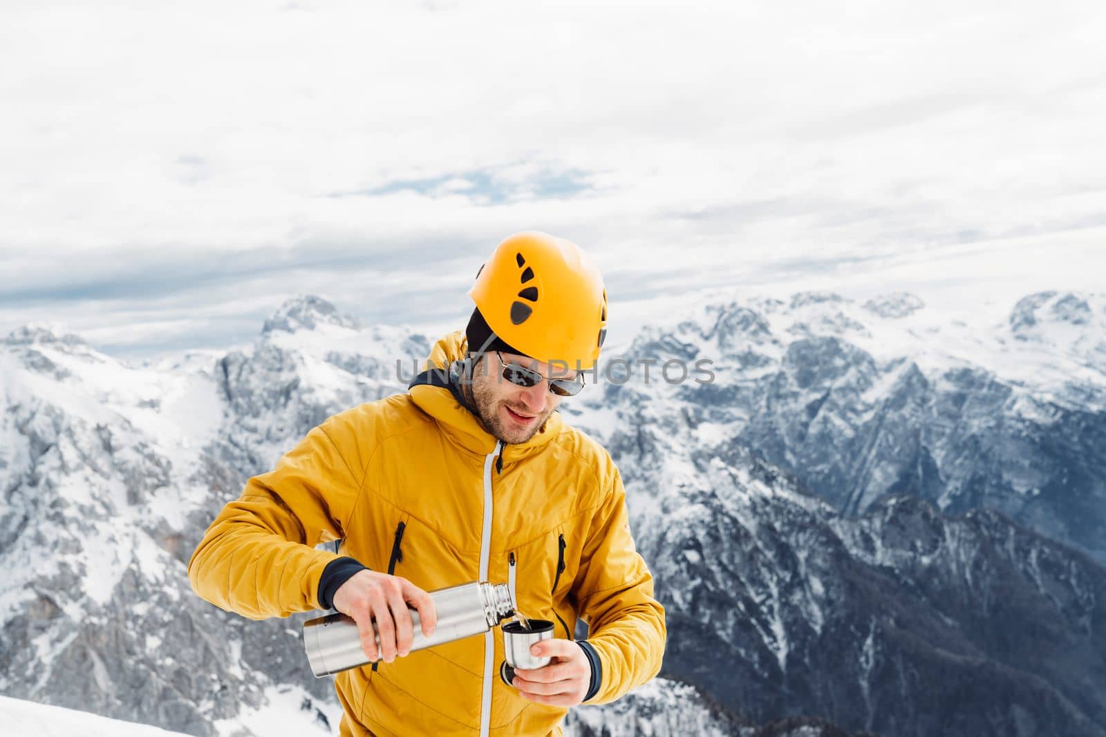 Mountaineer in a yellow jacket and helmet pouring himself some tea, view of snowy Alps in the background by VisualProductions