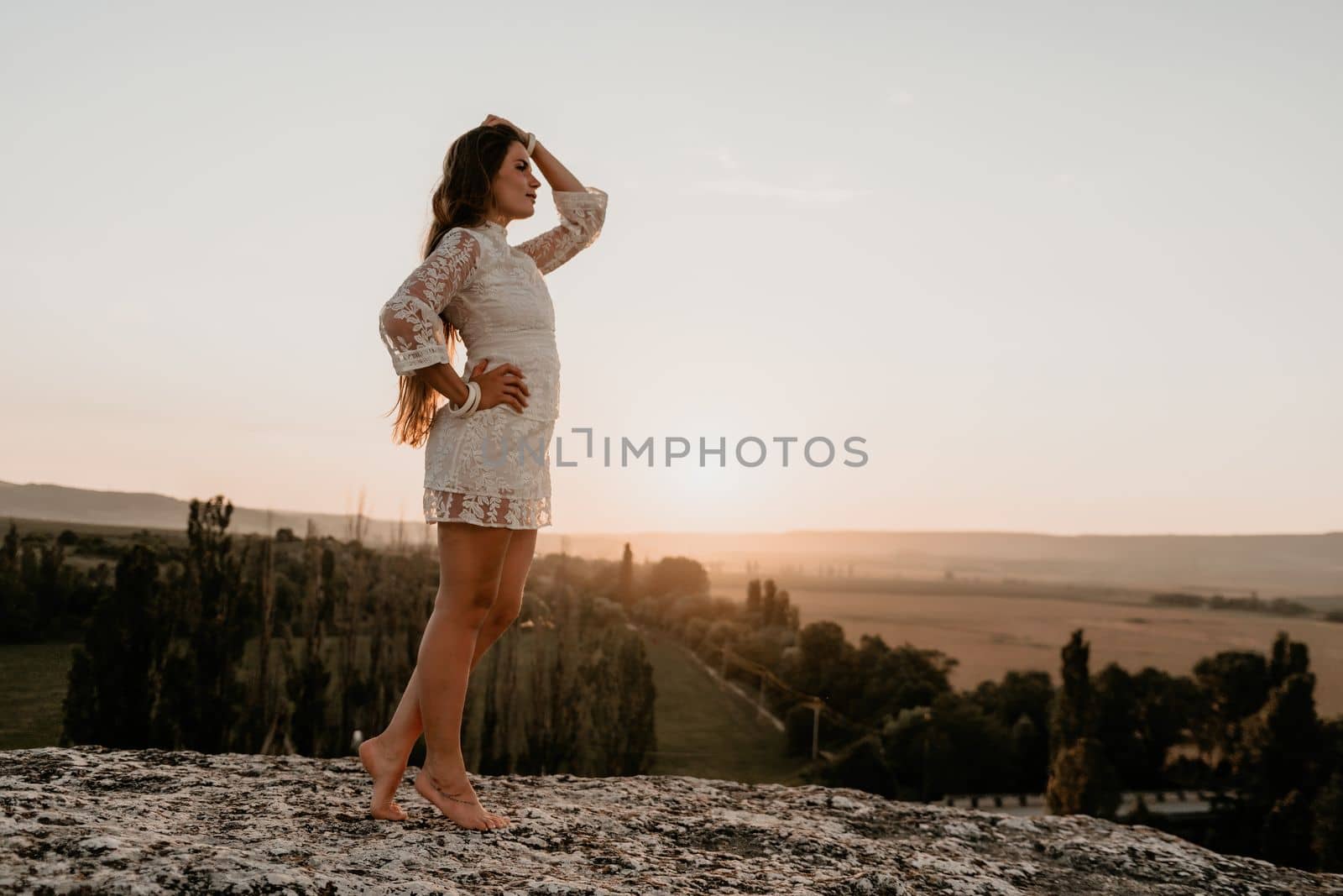 Happy woman in white boho dress on sunset in mountains. Romantic woman with long hair standing with her back on the sunset in nature in summer with open hands. Silhouette. Nature. Sunset. by panophotograph