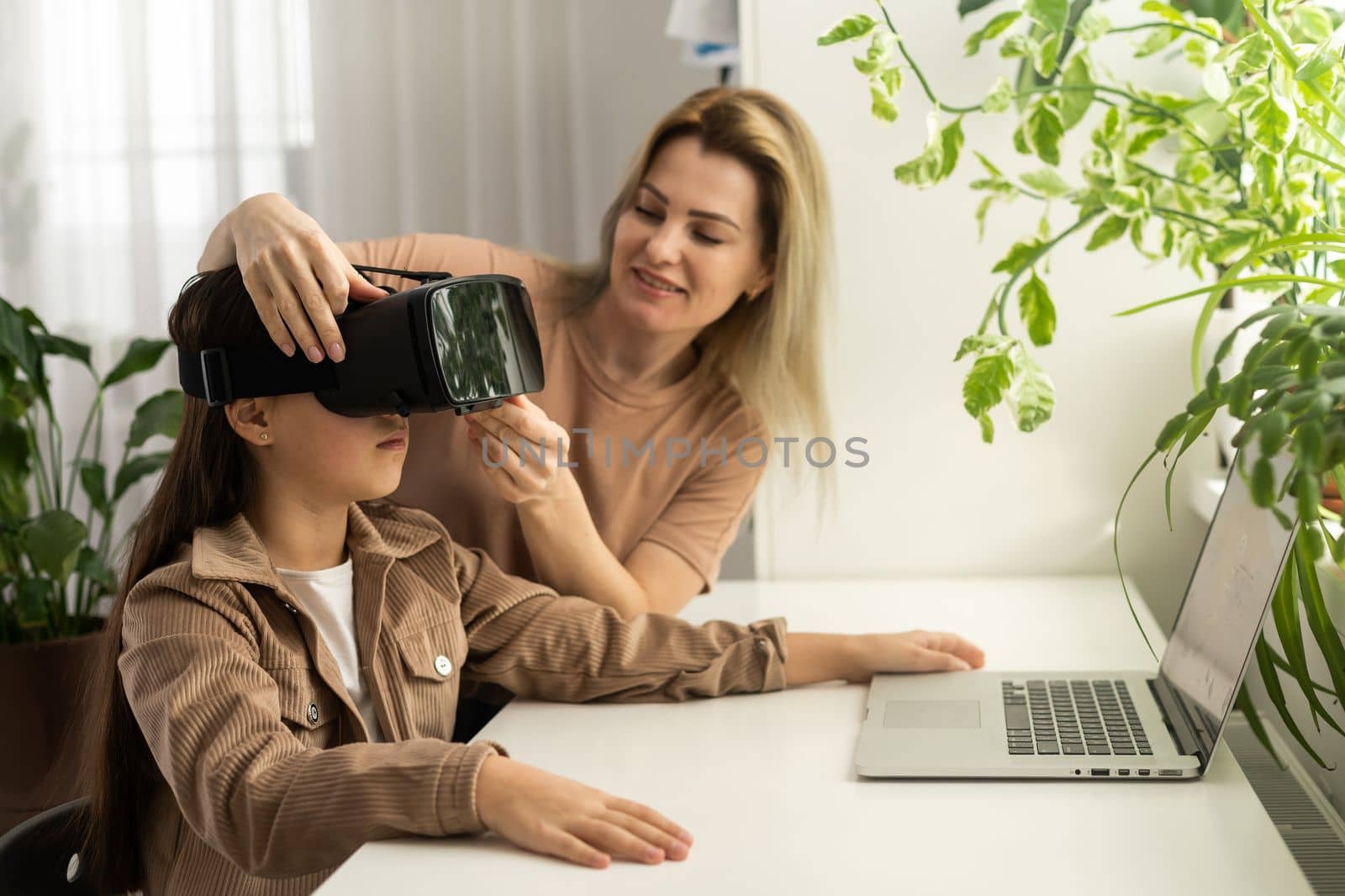 Blond excited mother helping her adorable girl teenager virtual using virtual reality headset at home interior. by Andelov13