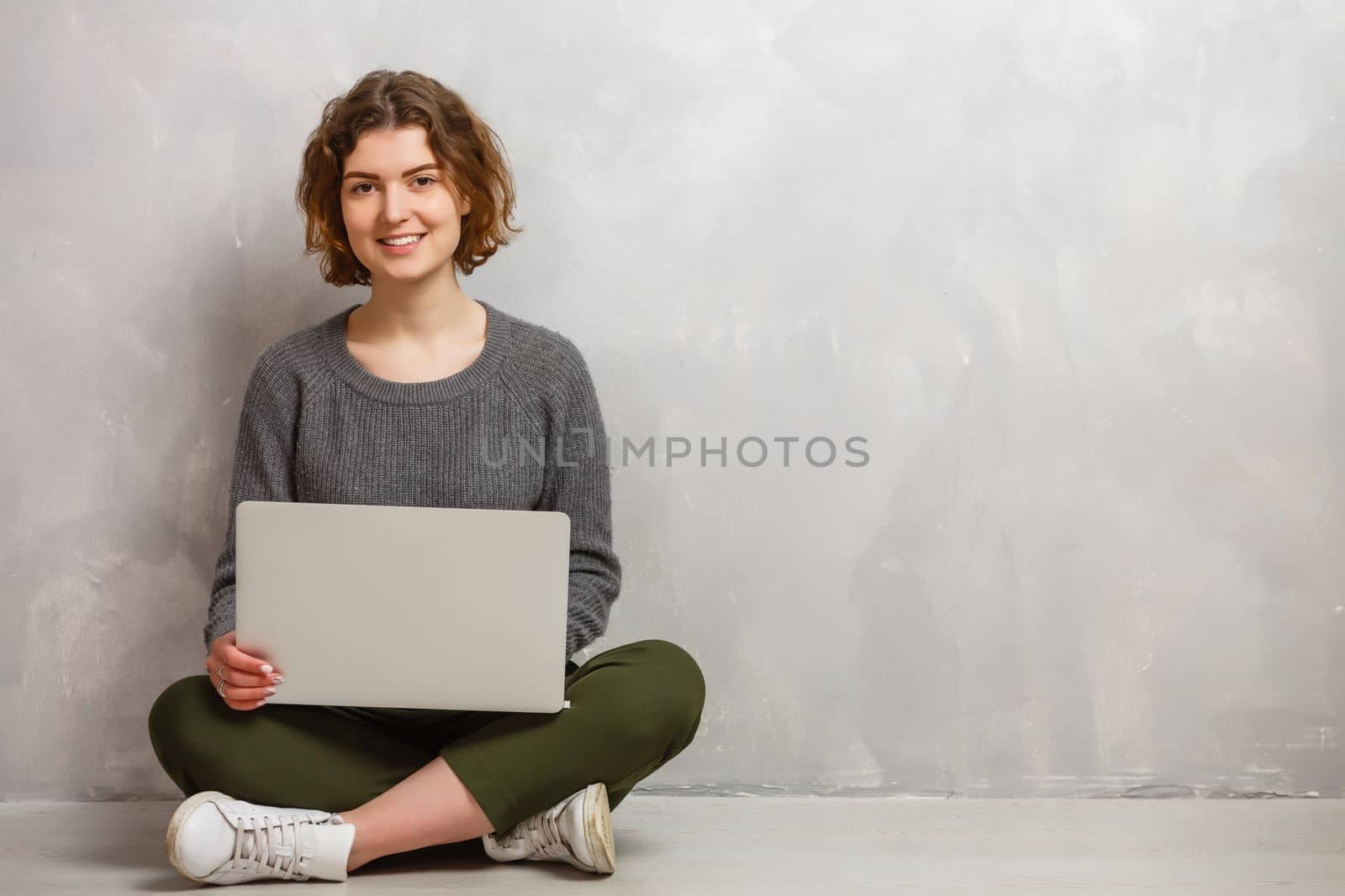 Happy young woman sitting on the floor with crossed legs and using laptop on gray background