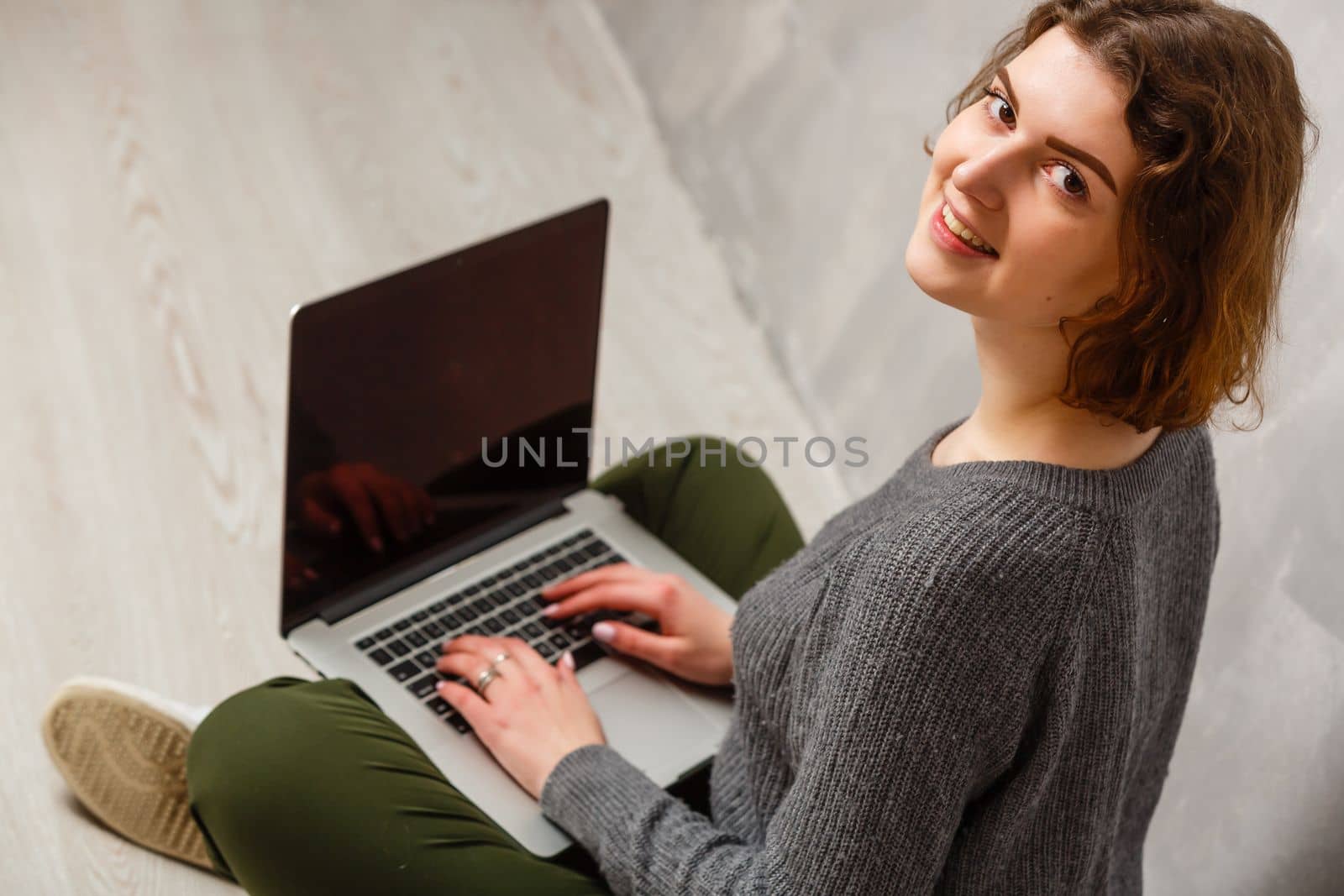 Portrait of satisfied female with beautiful smile enjoying watching movie in silver computer and sitting in lotus pose on the floor over grey wall
