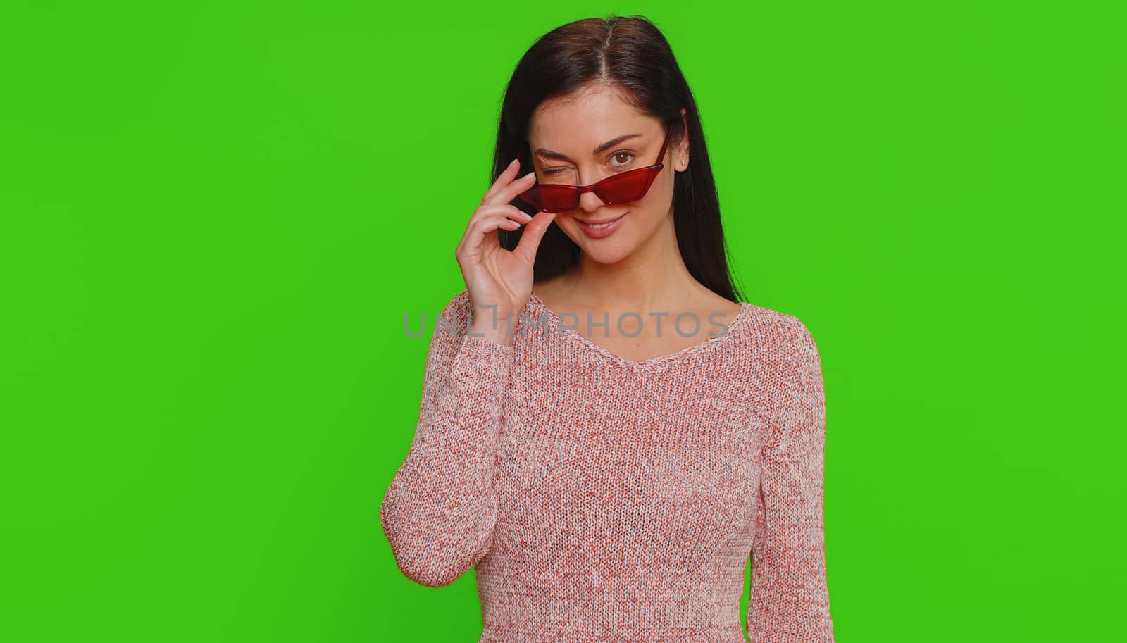Portrait of happy pretty young woman in sweater wearing sunglasses, looking at camera with toothy charming smile, flirting, expressing optimism. Millennial girl isolated on green chroma key background
