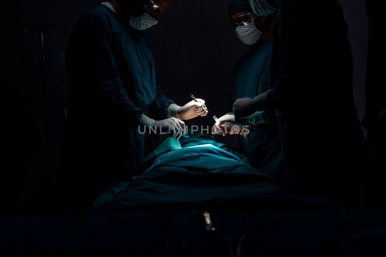 Surgical team performing surgery to patient in sterile operating room. In a surgery room lit by a lamp, a professional and confident surgical team provides medical care to an unconscious patient.