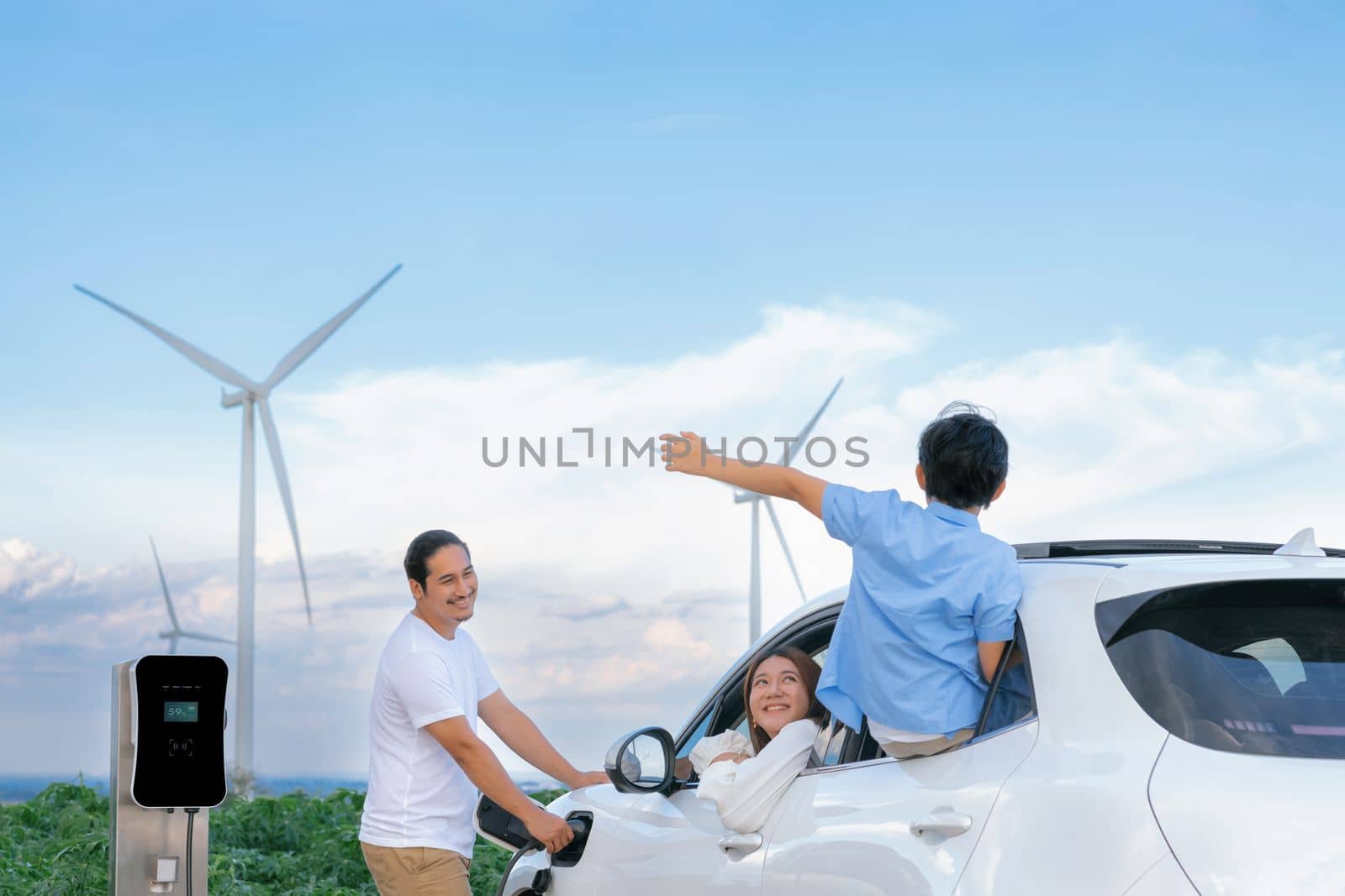 Concept of progressive happy family enjoying their time at wind farm with electric vehicle. Electric vehicle driven by clean renewable energy from wind turbine generator for charging station.