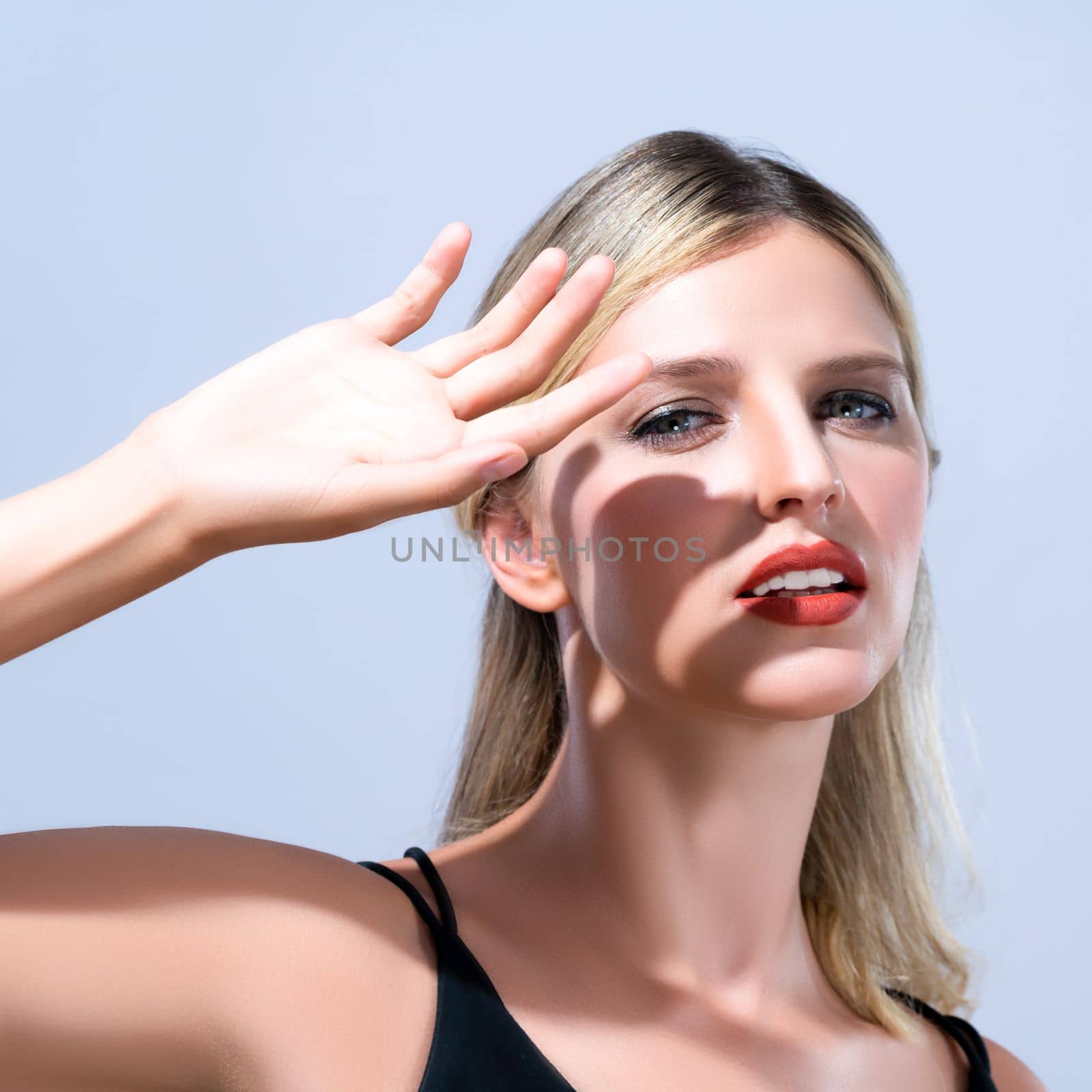 Closeup young blond hair woman with perfect skin and soft alluring facial makeup raise her hand cover her face from bright sunlight in isolated background for skincare sunscreen product.