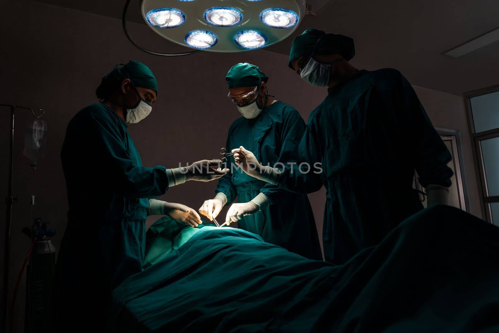 Surgical team performing surgery to patient in sterile operating room. In a surgery room lit by a lamp, a professional and confident surgical team provides medical care to an unconscious patient.