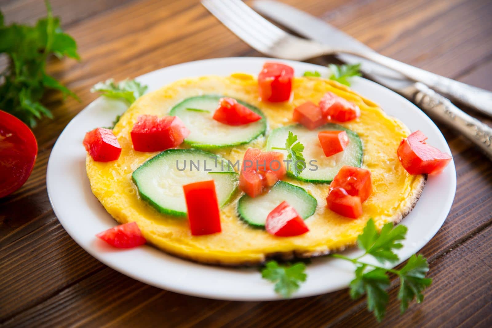 Fried omelet with zucchini, tomatoes, herbs in a plate on a wooden table.