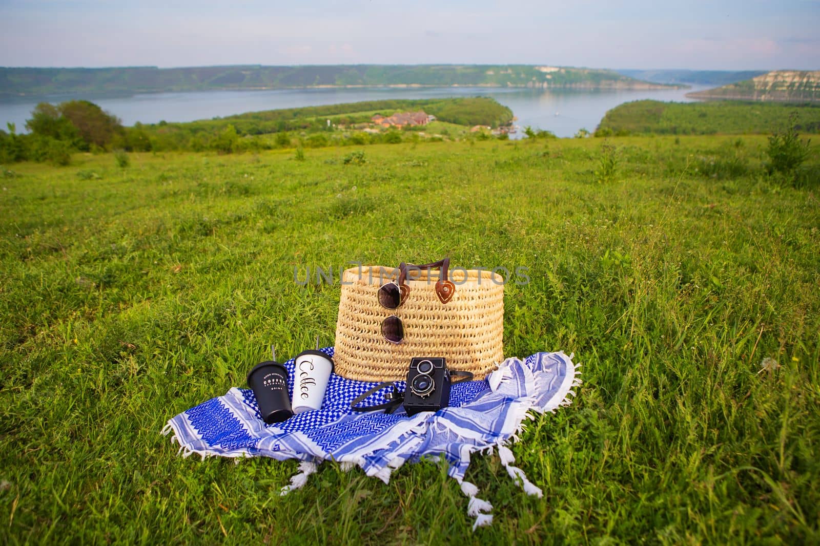 A very beautiful view of the lake. A straw picnic basket stands on a blue blanket on green grass along with glasses, a camera and coffee cups. Recreation in the open air. by sfinks