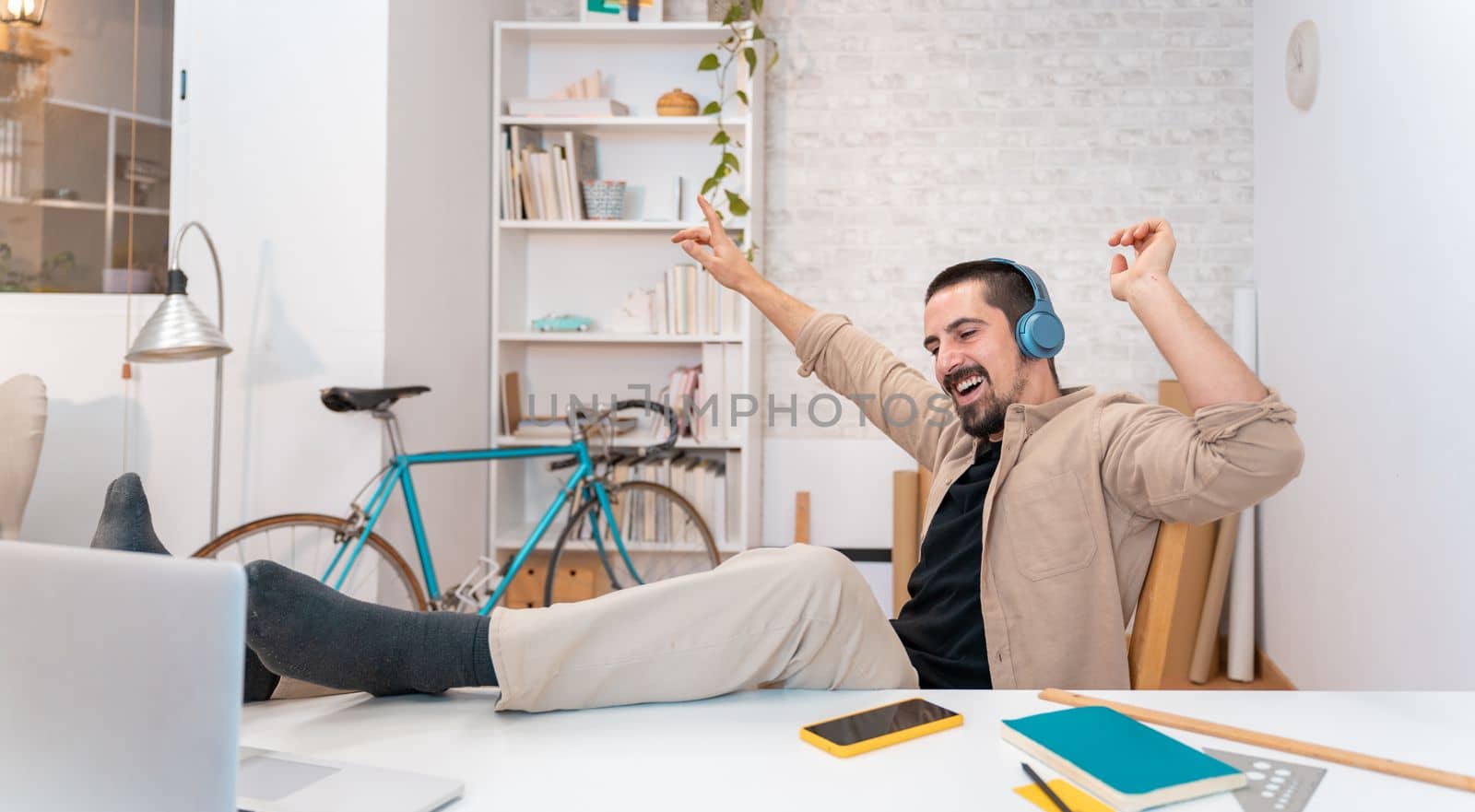 Happy smiling young man in headphones with laptop computer listening music and dancing in home office. Cheerful entrepreneur working from home.