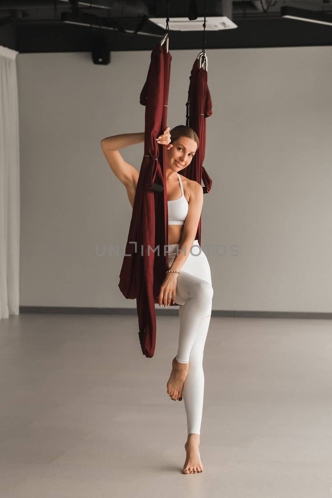 Portrait of a girl in white sportswear sitting in a hanging yoga hammock in the fitness room.
