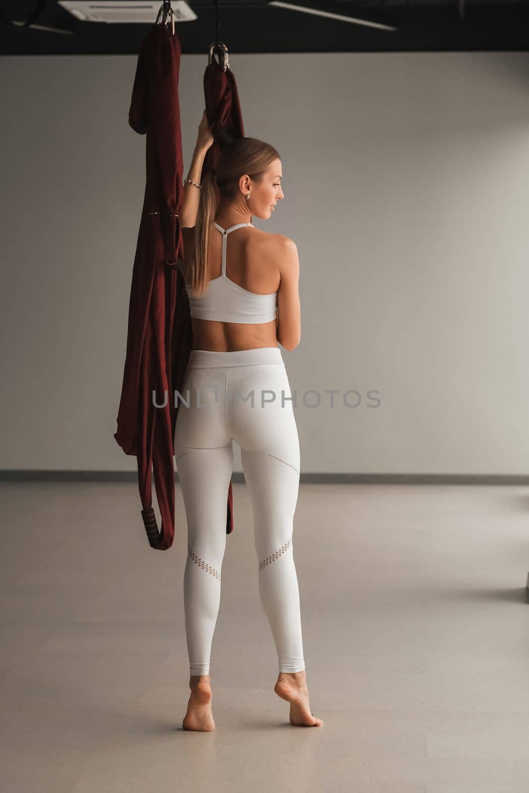 A portrait of a girl in white sportswear stands near a hanging Yoga hammock in the fitness room.