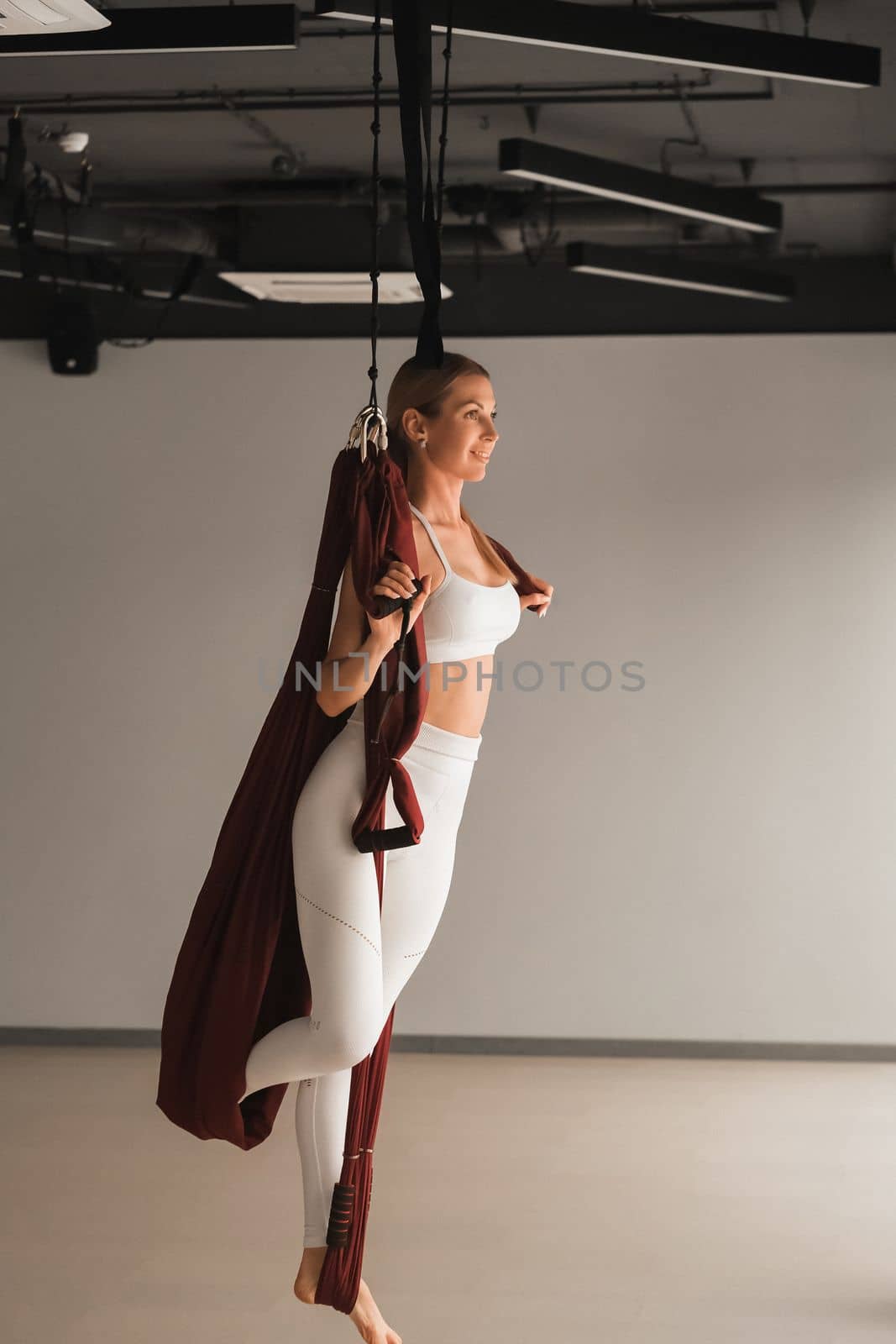 A girl in white sportswear does yoga on a hanging hammock in the fitness room.