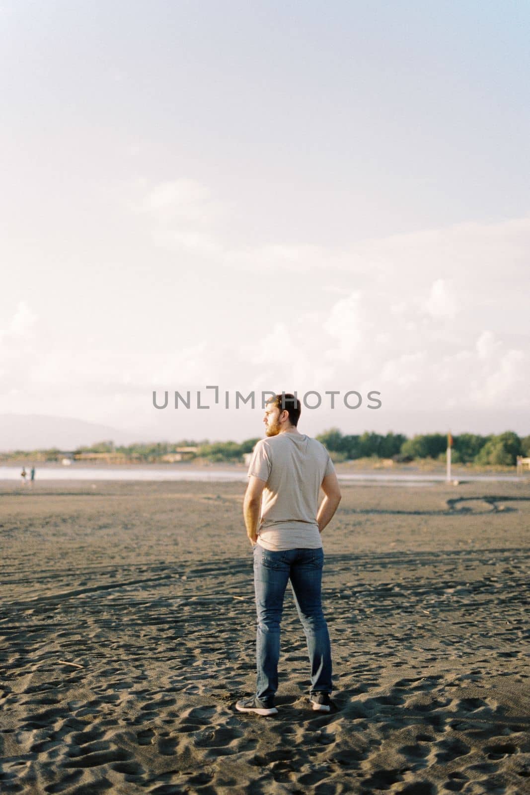 Young man in jeans stands on a sandy beach and looks at the sea. High quality photo