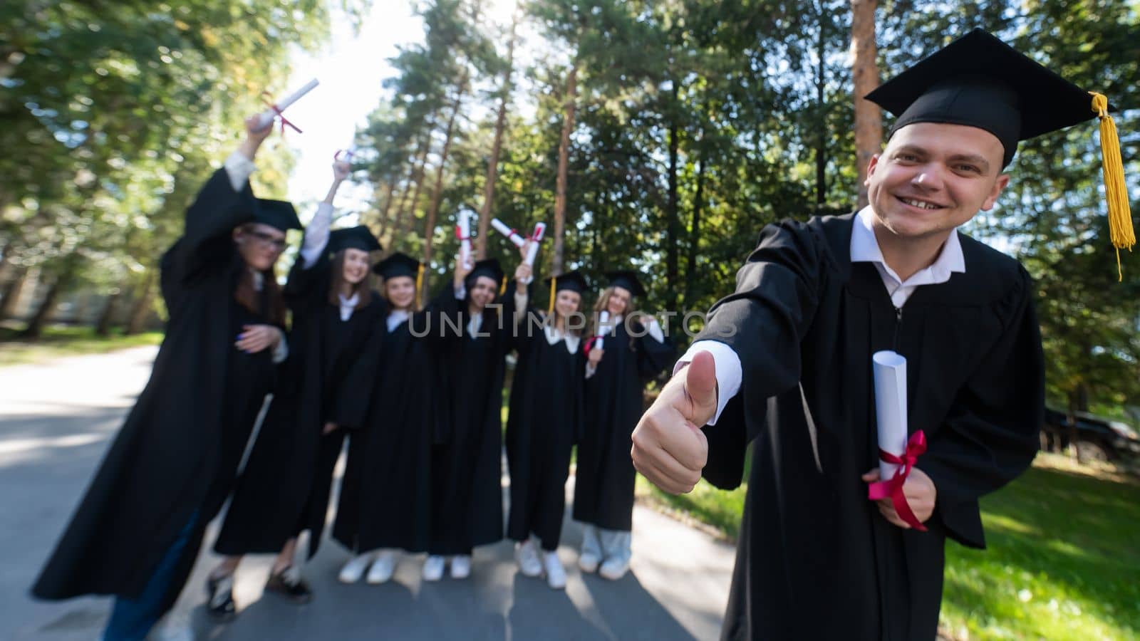 Happy young caucasian male graduate showing thumbs up. A group of graduate students outdoors. by mrwed54