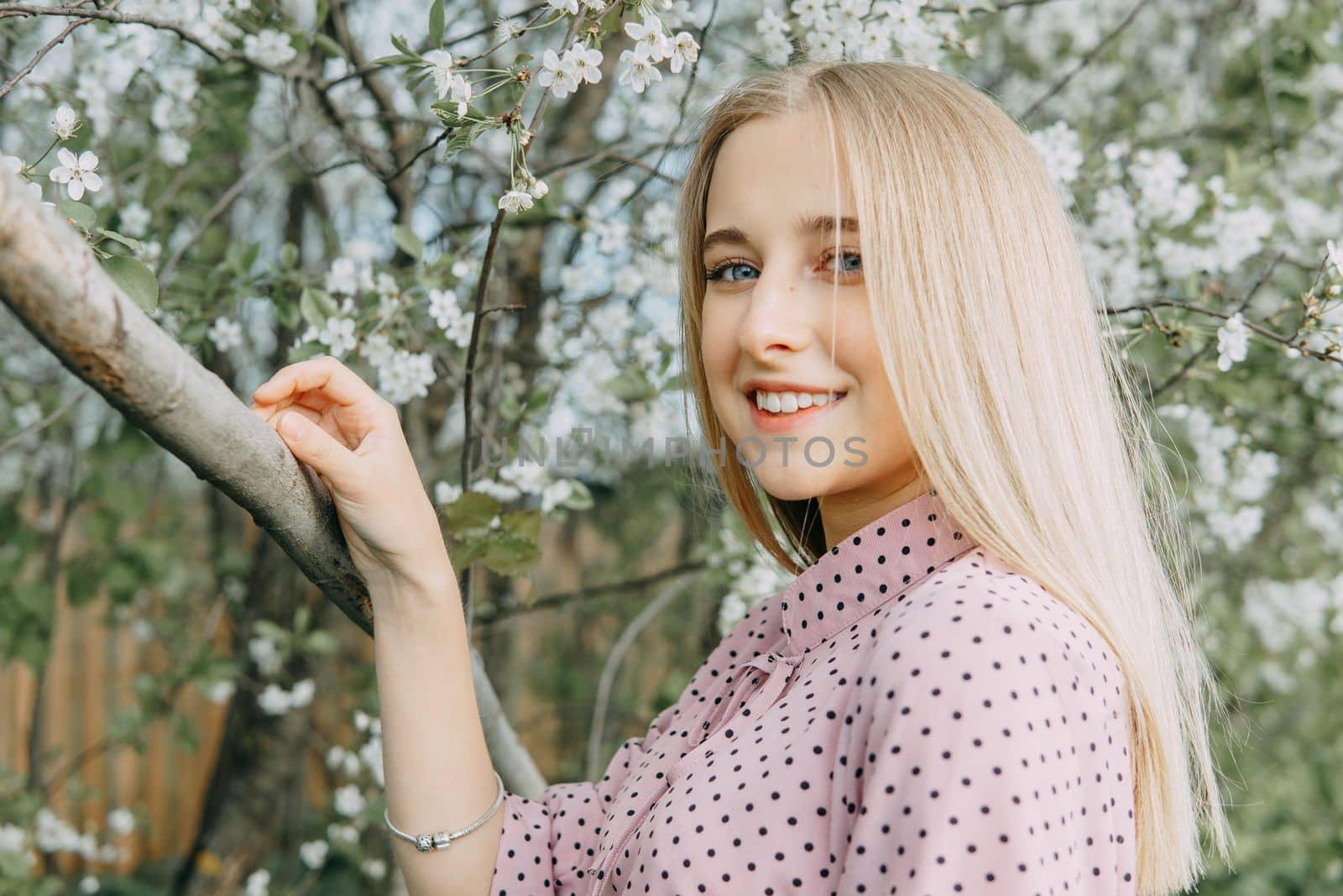 Blonde girl on a spring walk in the garden with cherry blossoms. Female portrait, close-up. A girl in a pink polka dot dress