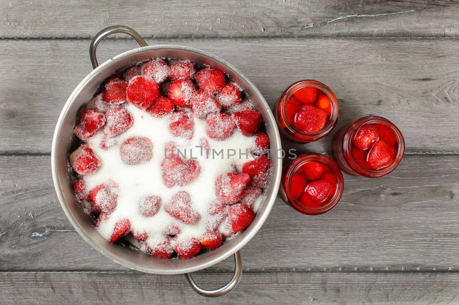 Tabletop view - steel put with strawberries covered in crystal sugar, three bottles with pickled strawberry next to it. Home made jam / compote preparation