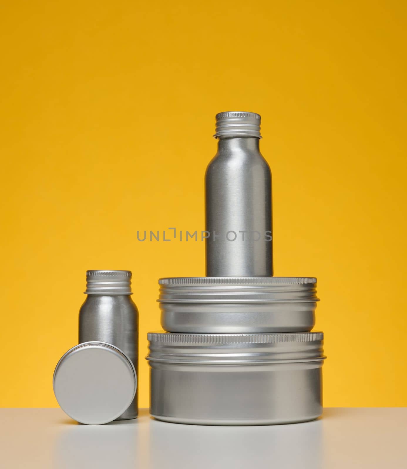 Empty silver metal bottle for cosmetics on white table, yellow background. Packaging for cream, gel, serum, advertising and product promotion