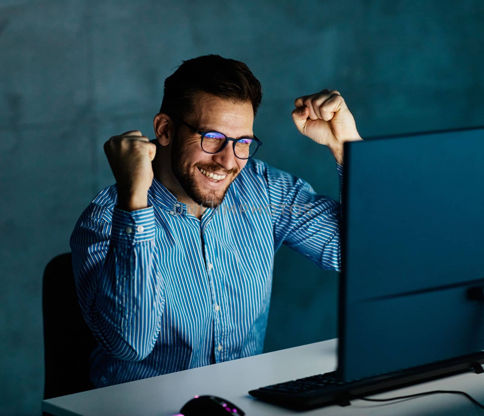 Portrait of a happy young businessman by monitor and desktop computer working late celebrating success and good news at night in office