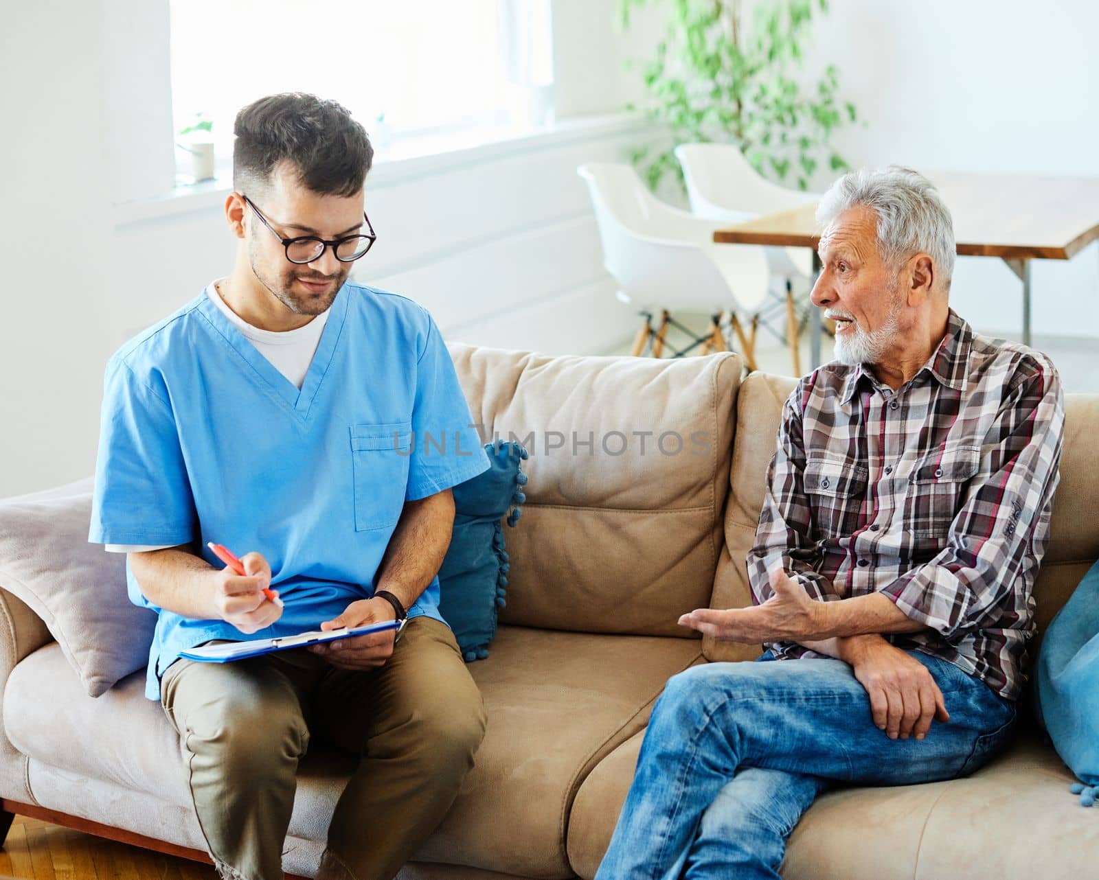 Doctor or nurse caregiver talking with senior man with chest pain and coughing problems on sofa at home or nursing home