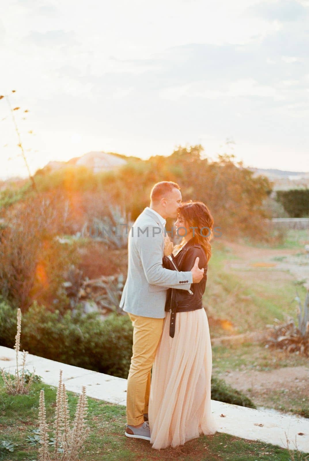 Groom kisses bride on the forehead standing on a stone path among the greenery. High quality photo