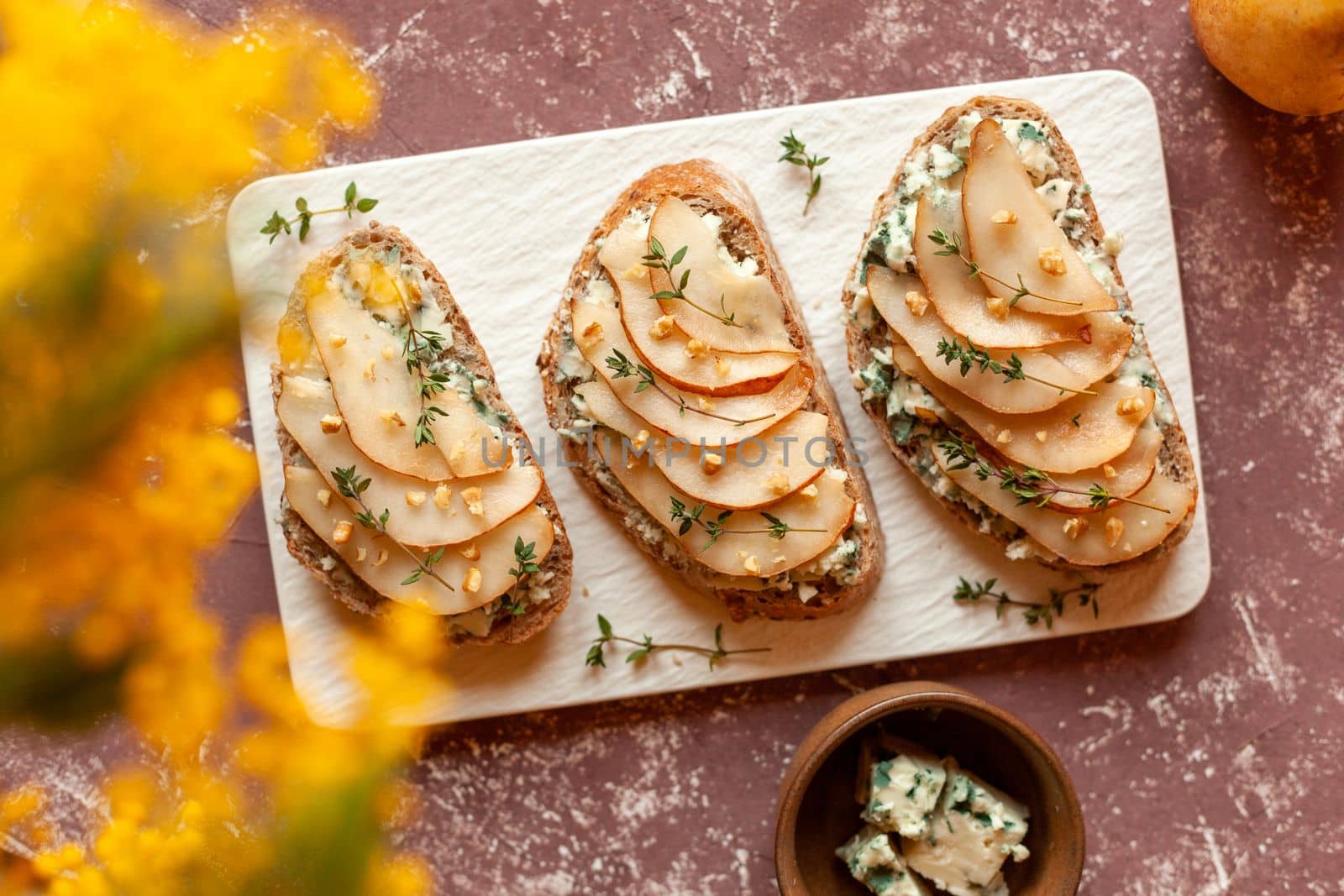 roquefort french cheese and sliced peaches toasts on a square plate, on a table under mimosa flowers, top view