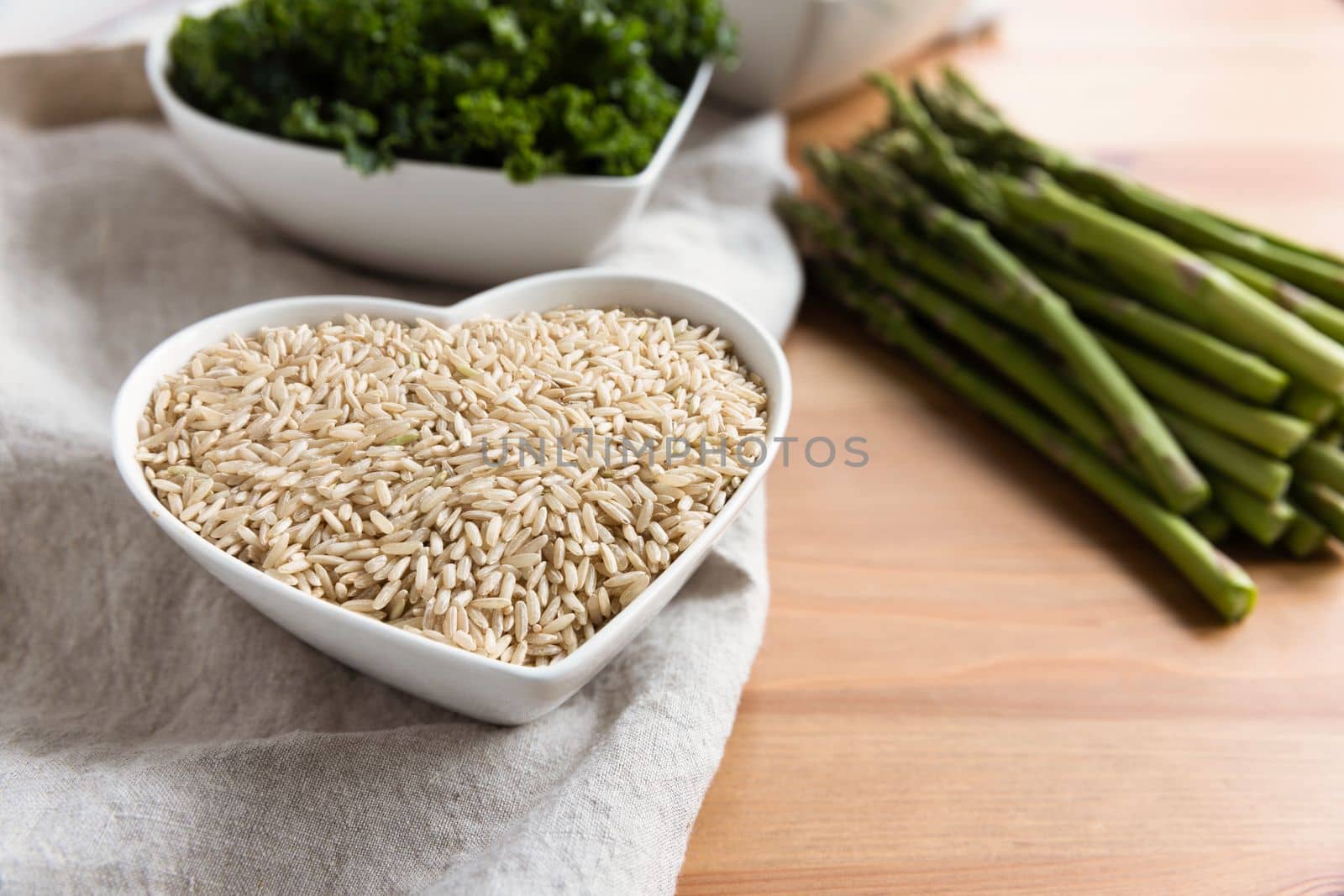 Healthy brown rice in a heart shaped bowl with asparagus and kale in the background, raw ingredients